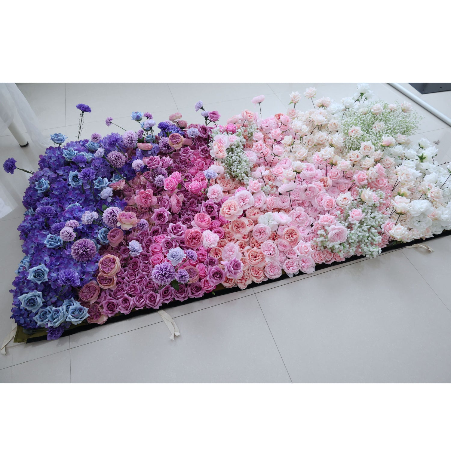 This photo showcases a large, colorful floral arrangement on a tiled floor. It features a gradient of blue, purple, pink, and white roses, hydrangeas, and baby&