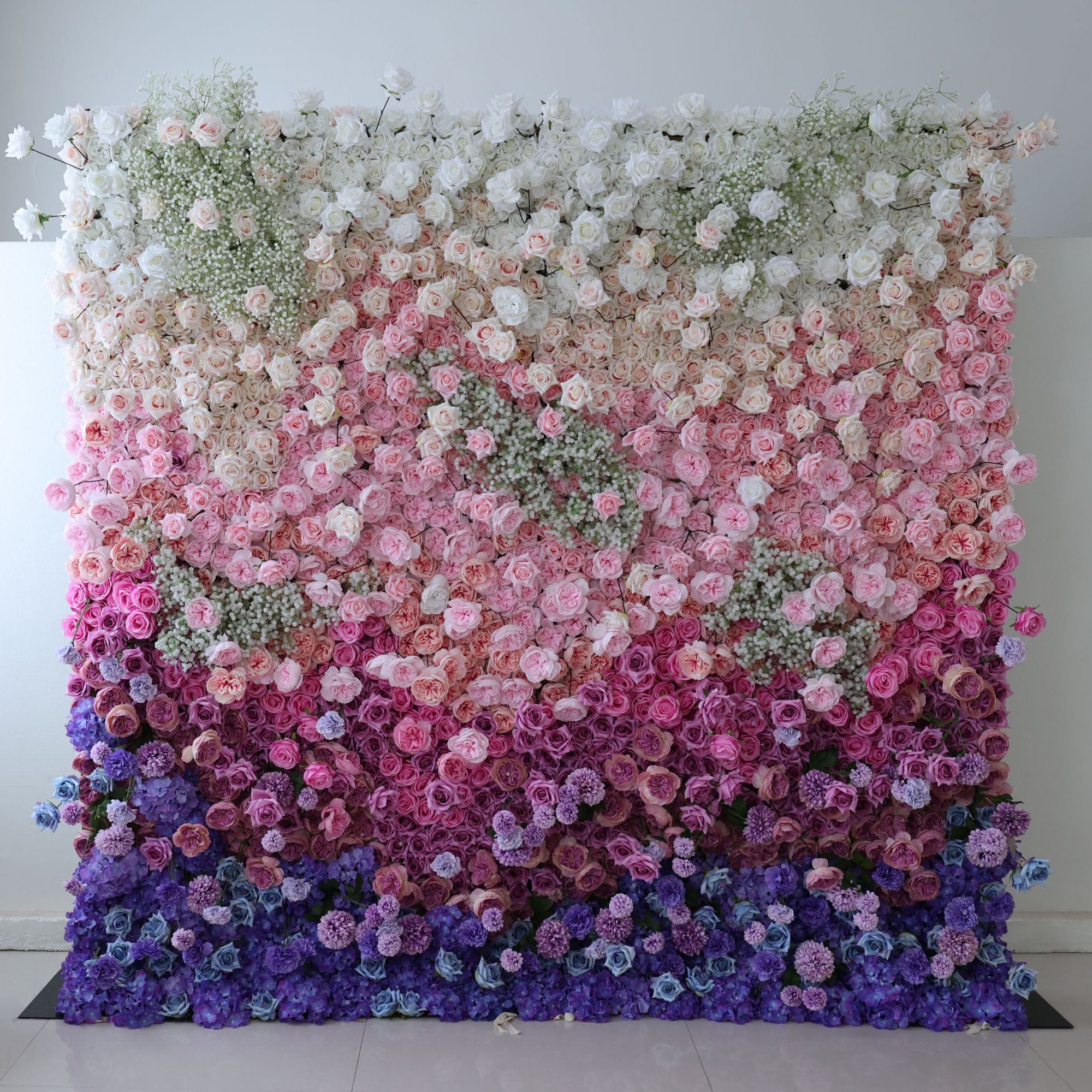 This photo features a lush floral wall with a gradient of white, pink, purple, and blue. Composed of roses, hydrangeas, and baby&