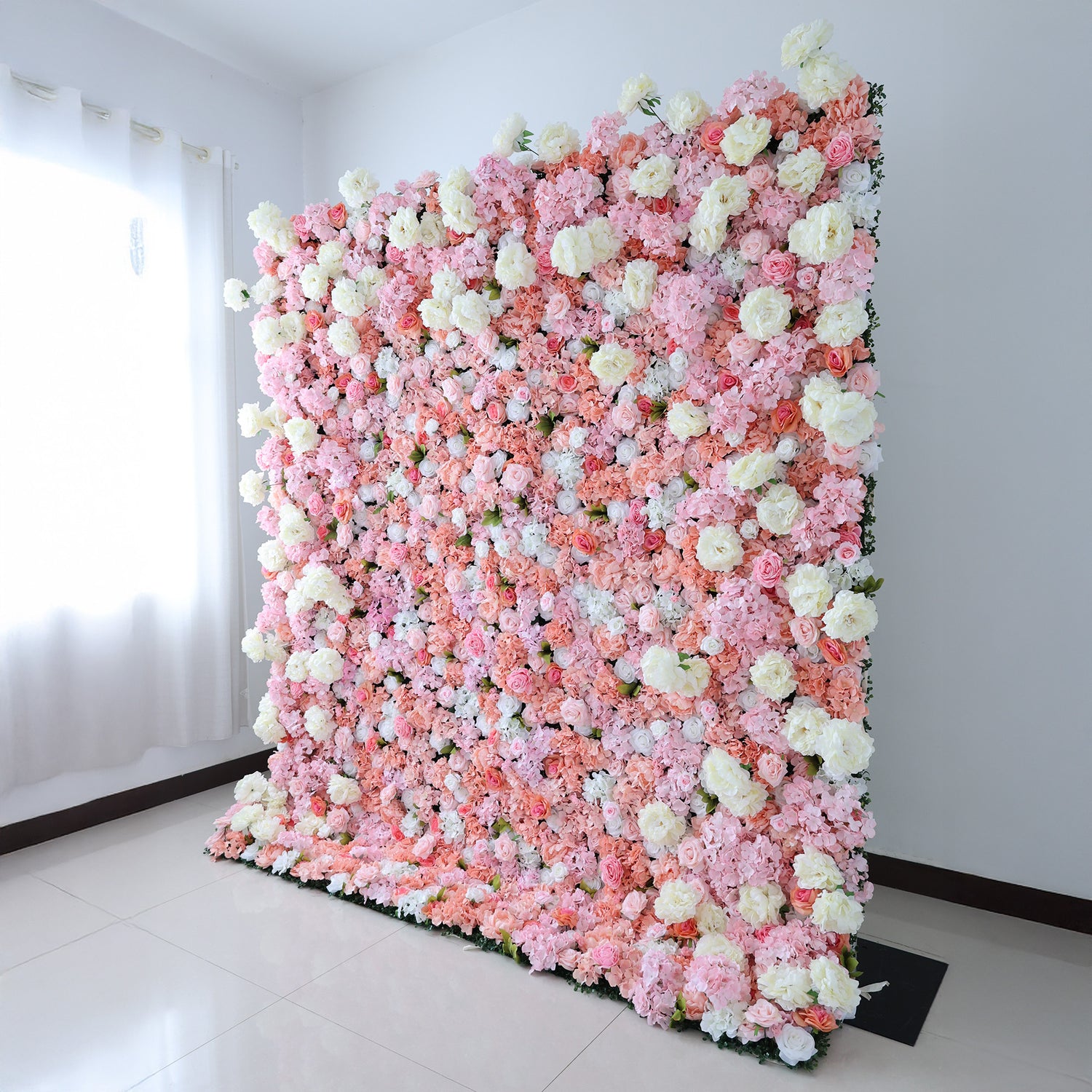 This image showcases a lush floral backdrop by ValarFlowers, set indoors. It features a dense arrangement of artificial flowers in shades of pink, peach, and white, creating a luxurious, textured display. Natural light from a window enhances the scene, exuding elegance and sophistication, perfect for adding a nature-inspired element to events or interiors.