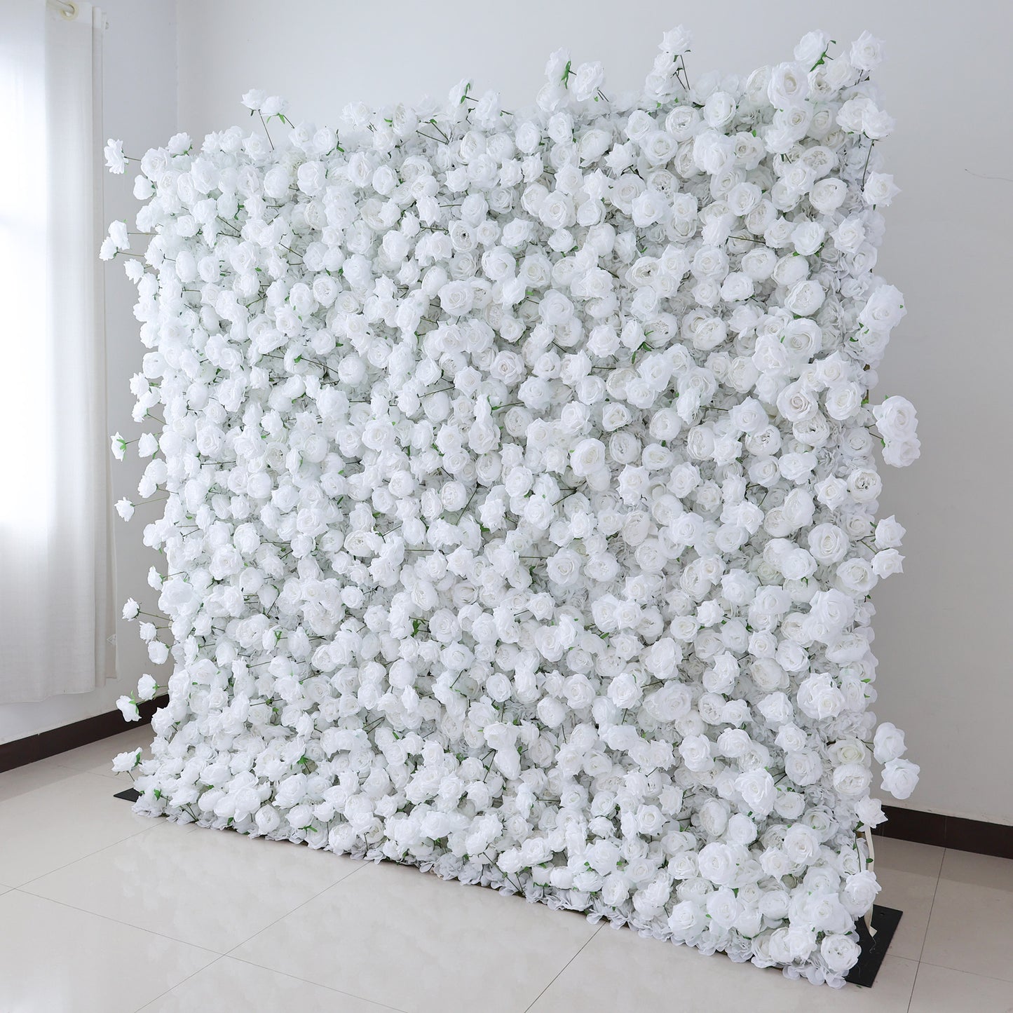 This image features a large, square floral wall indoors. It's densely covered with pure white artificial roses, creating a clean, elegant, and serene display. The flowers are tightly packed, giving a sense of opulence and purity. Set against a plain white wall with a shiny tiled floor, and with a curtain nearby, the scene exudes an atmosphere of sophistication and natural beauty, perfect for decorative purposes in events or as a striking interior backdrop.