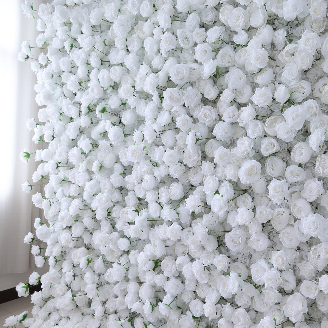 This image presents a dense arrangement of white artificial roses, creating a lush and voluminous floral display. The flowers are tightly packed, with their soft, creamy petals and subtle green stems contributing to a sense of purity and elegance. Set indoors, likely near a window with light filtering through, the scene exudes a serene and sophisticated atmosphere, ideal for decorative purposes in events or as a striking interior backdrop, evoking feelings of natural beauty and refinement.