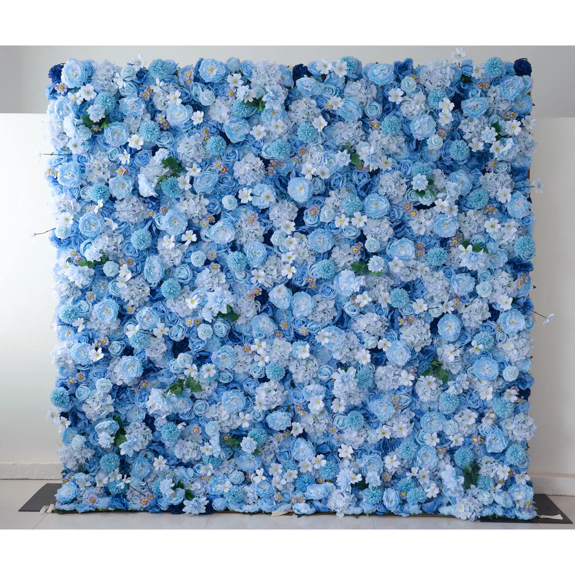 A large artificial flower wall features a lush arrangement of blue blooms, including roses and hydrangeas, interspersed with tiny white flowers and green foliage. The varying shades of blue create a harmonious, serene backdrop, perfect for adding an elegant, calming touch to events or decor. 