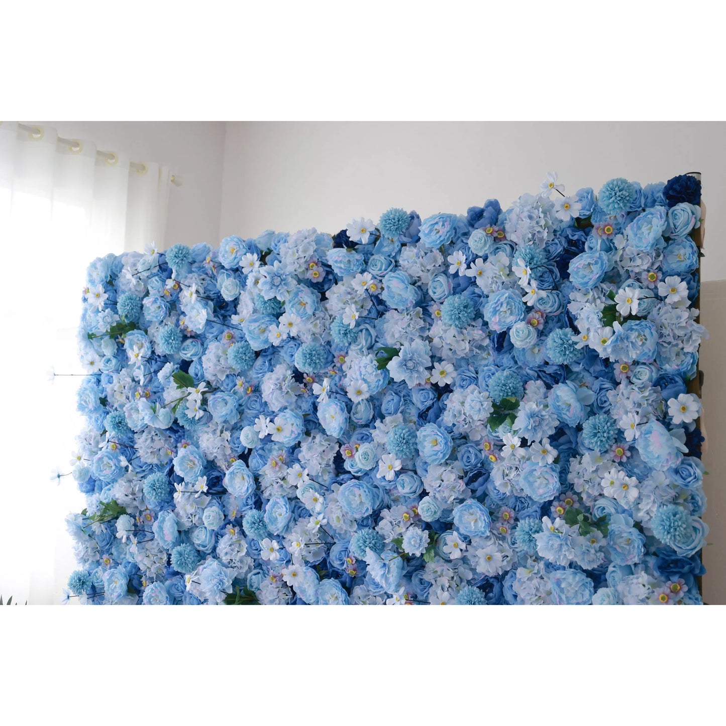 A large artificial flower wall features a lush mix of blue blooms—roses, hydrangeas, and delicate white flowers with yellow centers. The varying shades of blue, from soft pastels to deeper tones, create a harmonious, serene backdrop. Green foliage adds a natural touch, making it perfect for elegant decor or event backdrops. 