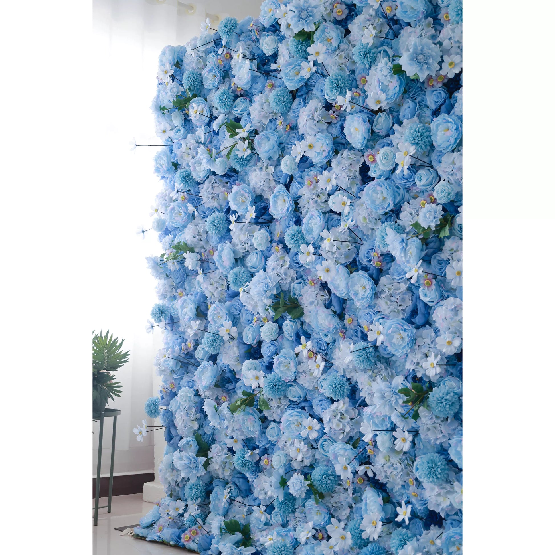A large artificial flower wall features a dense arrangement of blue blooms, including roses, hydrangeas, and small white flowers with yellow centers. The varying shades of blue create a harmonious, serene backdrop, enhanced by green foliage. Perfect for adding an elegant, calming touch to events or decor, it exudes a lush, romantic charm. 