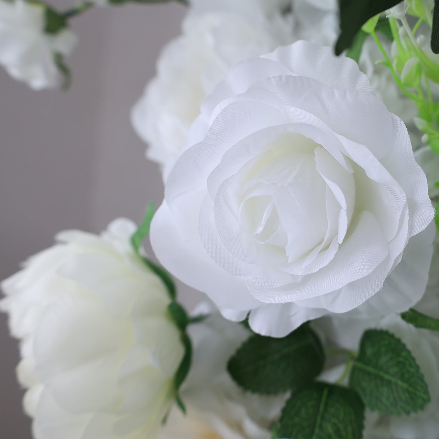 This image showcases a beautiful artificial white rose by Valar Flowers. The rose features delicate, layered petals with a soft, realistic texture. Surrounding it are other white blooms and lush green foliage, creating a cohesive and elegant floral arrangement. Perfect for adding a touch of timeless beauty to events or spaces, this piece exudes a refined and sophisticated charm, characteristic of ValarFlowers' craftsmanship.