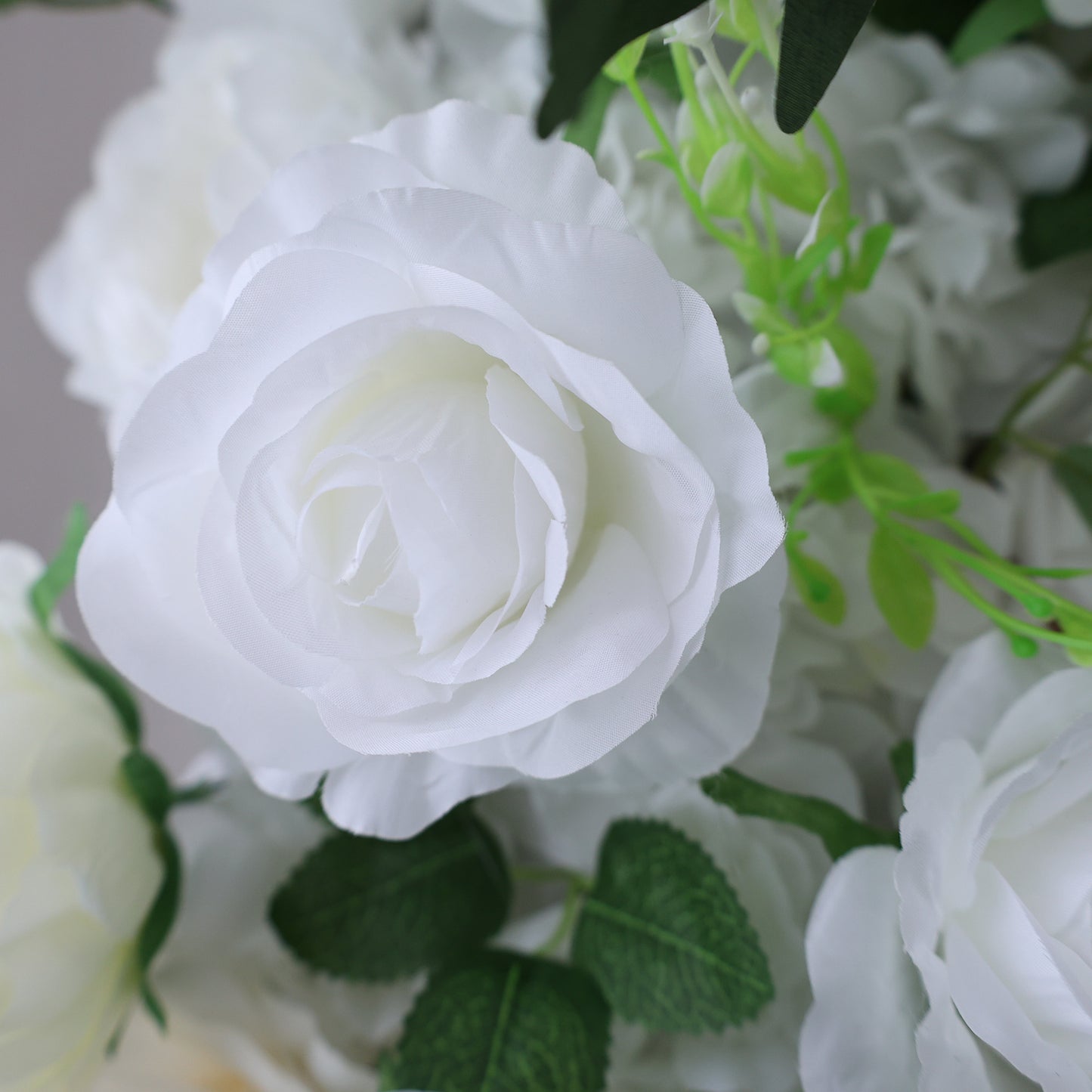 This image showcases a beautiful artificial white rose by Valar Flowers. The rose features delicate, layered petals with a soft, realistic texture. Surrounding it are other white blooms and lush green foliage, creating a cohesive and elegant floral arrangement. Perfect for adding a touch of timeless beauty to events or spaces, this piece exudes a refined and sophisticated charm, characteristic of Valar Flowers' craftsmanship.