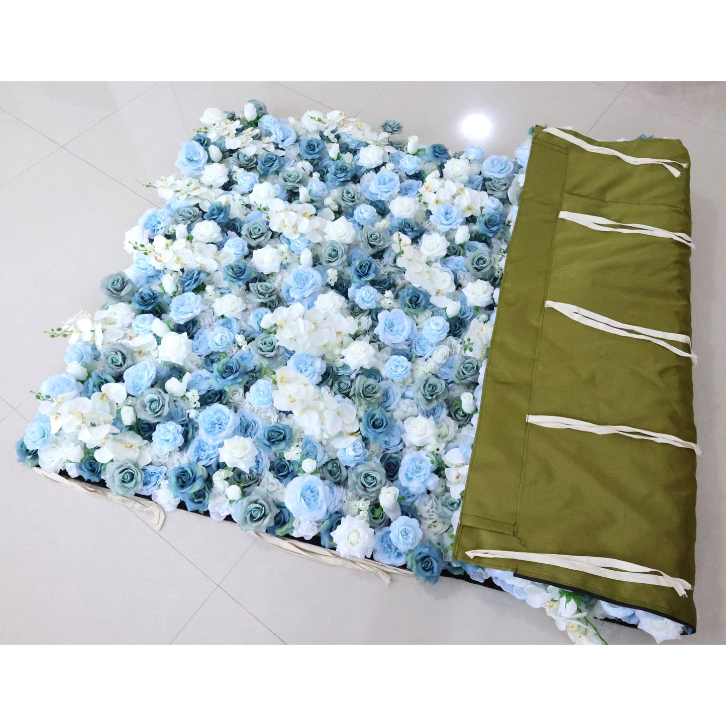 This photo shows a floral backdrop laid out on the floor. The backdrop is densely covered with blue and white roses and white orchids, creating a lush and elegant appearance. To the right, there's a green fabric with white ties, likely used for storage or transport. This setup is ideal for events like weddings or galas, offering a sophisticated and visually appealing decorative element. 