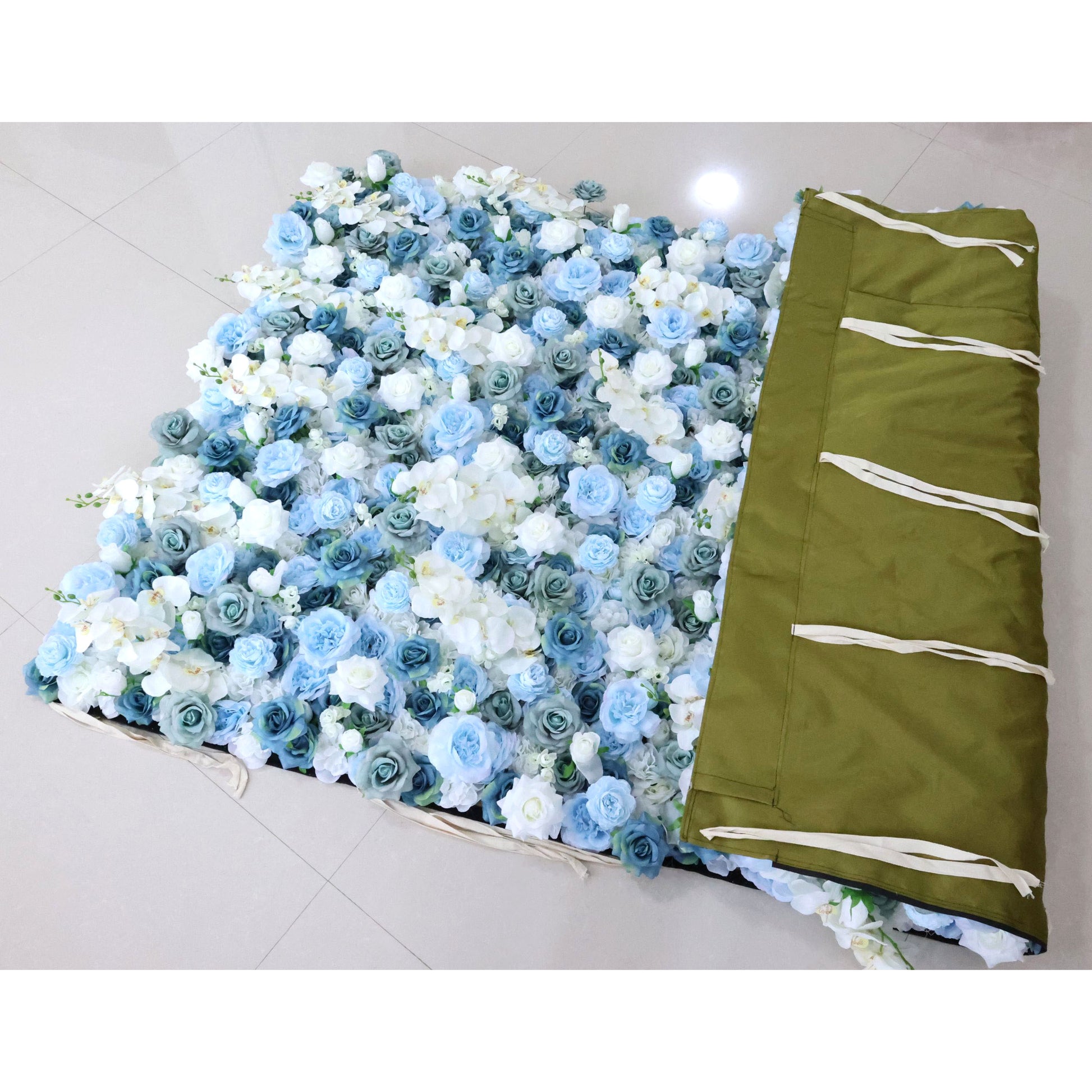This photo shows a floral backdrop laid out on the floor. The backdrop is densely covered with blue and white roses and white orchids, creating a lush and elegant appearance. To the right, there's a green fabric with white ties, likely used for storage or transport. This setup is ideal for events like weddings or galas, offering a sophisticated and visually appealing decorative element. 