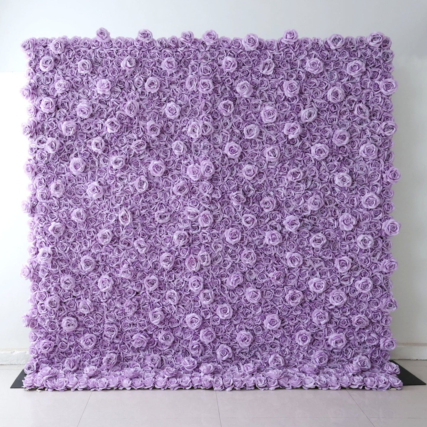 A square Valar Flowers floral wall panel, fully assembled against a plain white wall on light tile flooring, is entirely covered in dense, uniform lavender fabric roses. The seamless, textured design delivers a sleek, monochromatic aesthetic, perfect for minimalist weddings, baby showers, or purple-themed event photo backdrops.