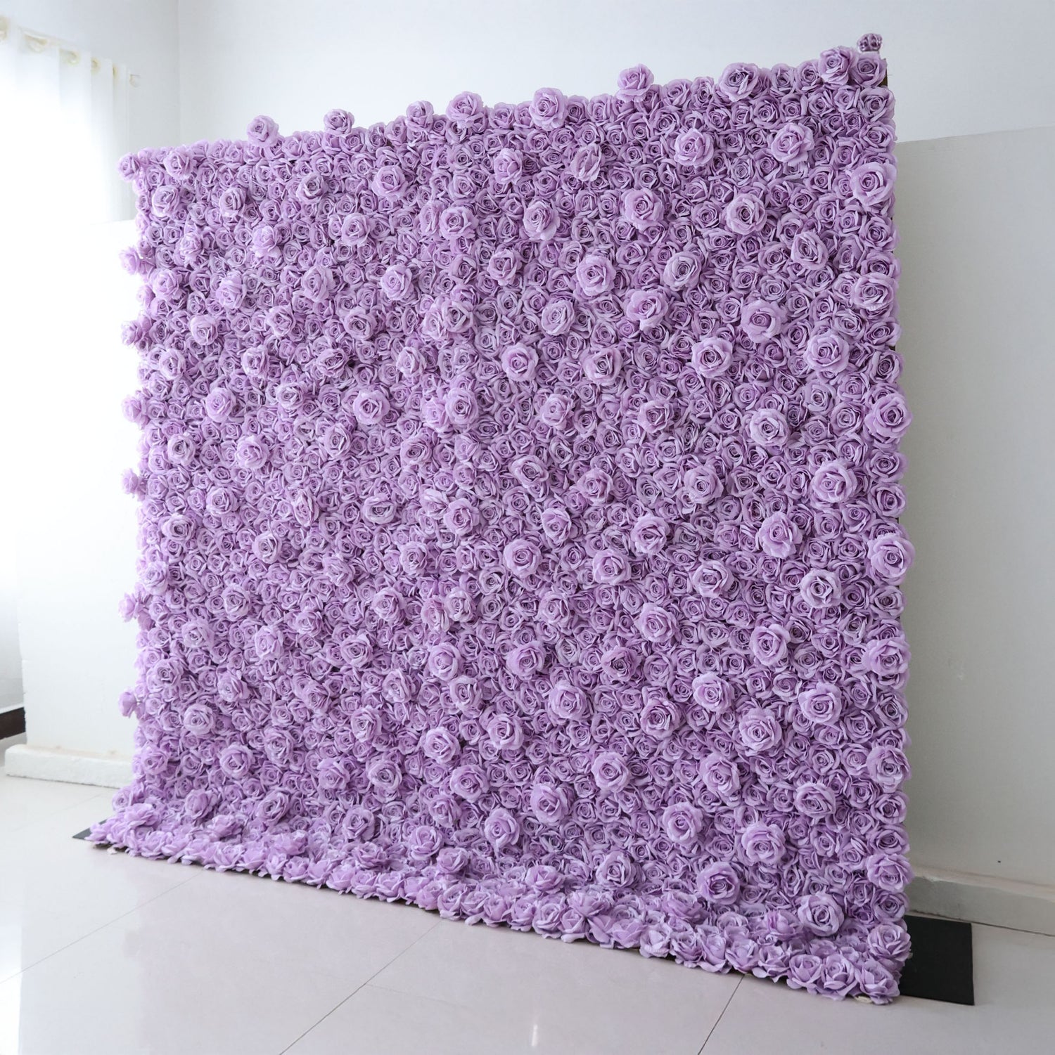 A tall, square Valar Flowers floral wall panel leans against a clean white wall on light tile flooring. It is densely covered in uniform, soft lavender fabric roses, creating a seamless, textured monochromatic surface. This sleek, minimalist design is ideal for wedding backdrops, purple-themed parties, or elegant photo booth setups.