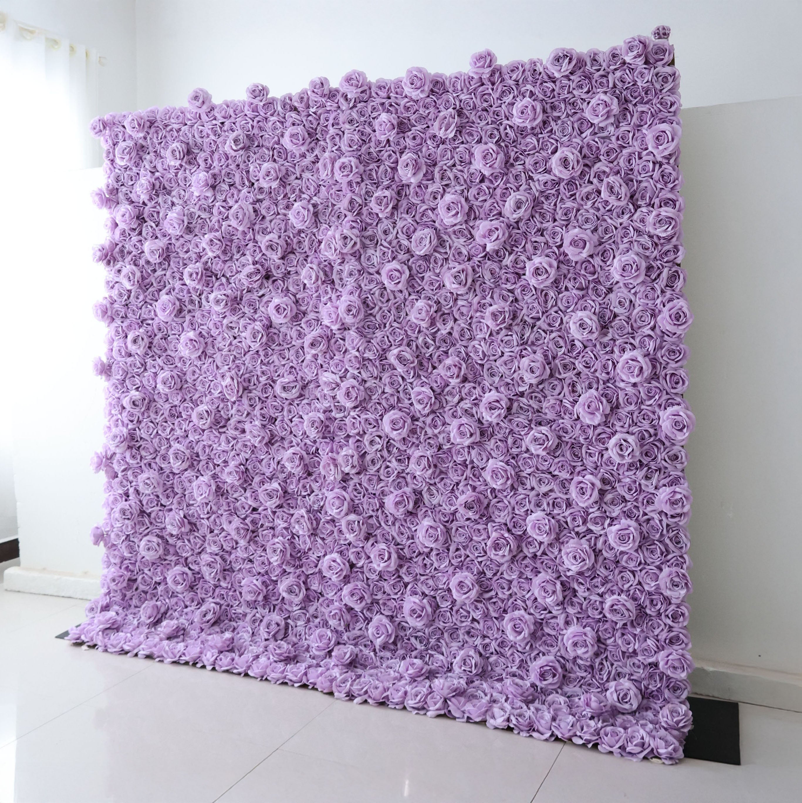 A tall, square Valar Flowers floral wall panel leans against a clean white wall on light tile flooring. It is densely covered in uniform, soft lavender fabric roses, creating a seamless, textured monochromatic surface. This sleek, minimalist design is ideal for wedding backdrops, purple-themed parties, or elegant photo booth setups.