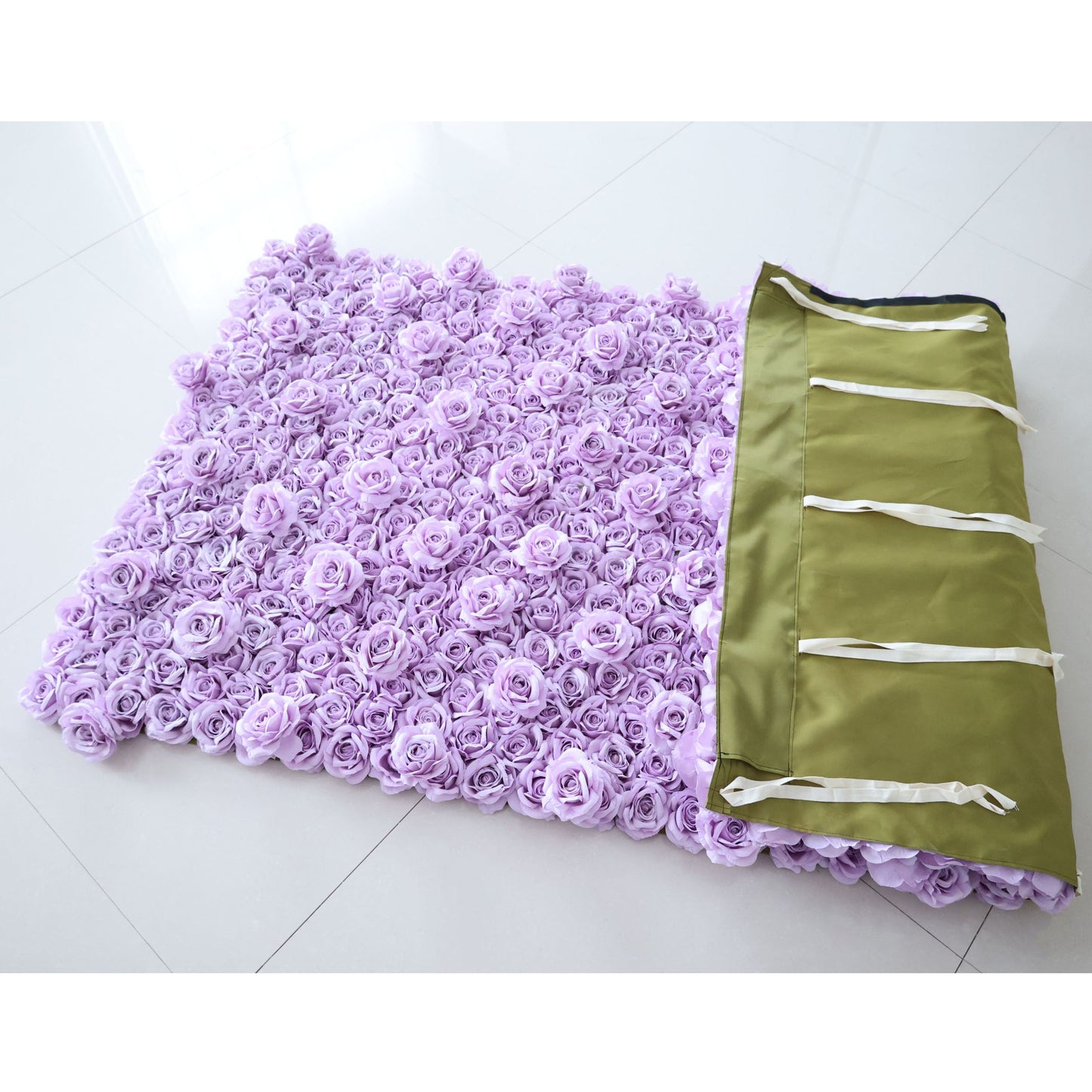 A flat-laid Valar Flowers lavender rose floral wall panel rests on a glossy white tiled floor, partially folded to reveal its olive-green backing with white hanging ribbons. Densely covered in soft, uniform lavender fabric roses, it highlights the product’s practical, easy-to-install design—ideal for quick wedding, party, or photo booth setup.