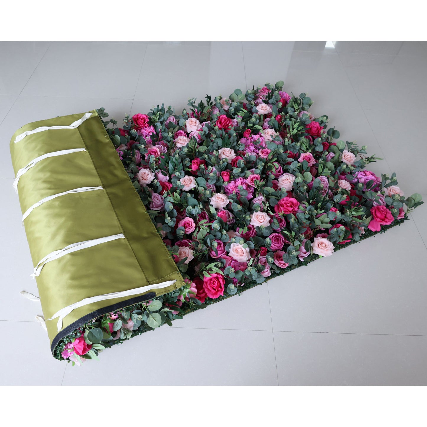 This image shows a 4x8ft roll of artificial floral mat. Partially unrolled, it reveals a dense arrangement of roses in shades of pink, red, and purple, interspersed with green eucalyptus leaves. The mat is edged with an olive - green fabric featuring white straps, lying on a smooth, light - colored tiled floor, ideal for decorative use in events.