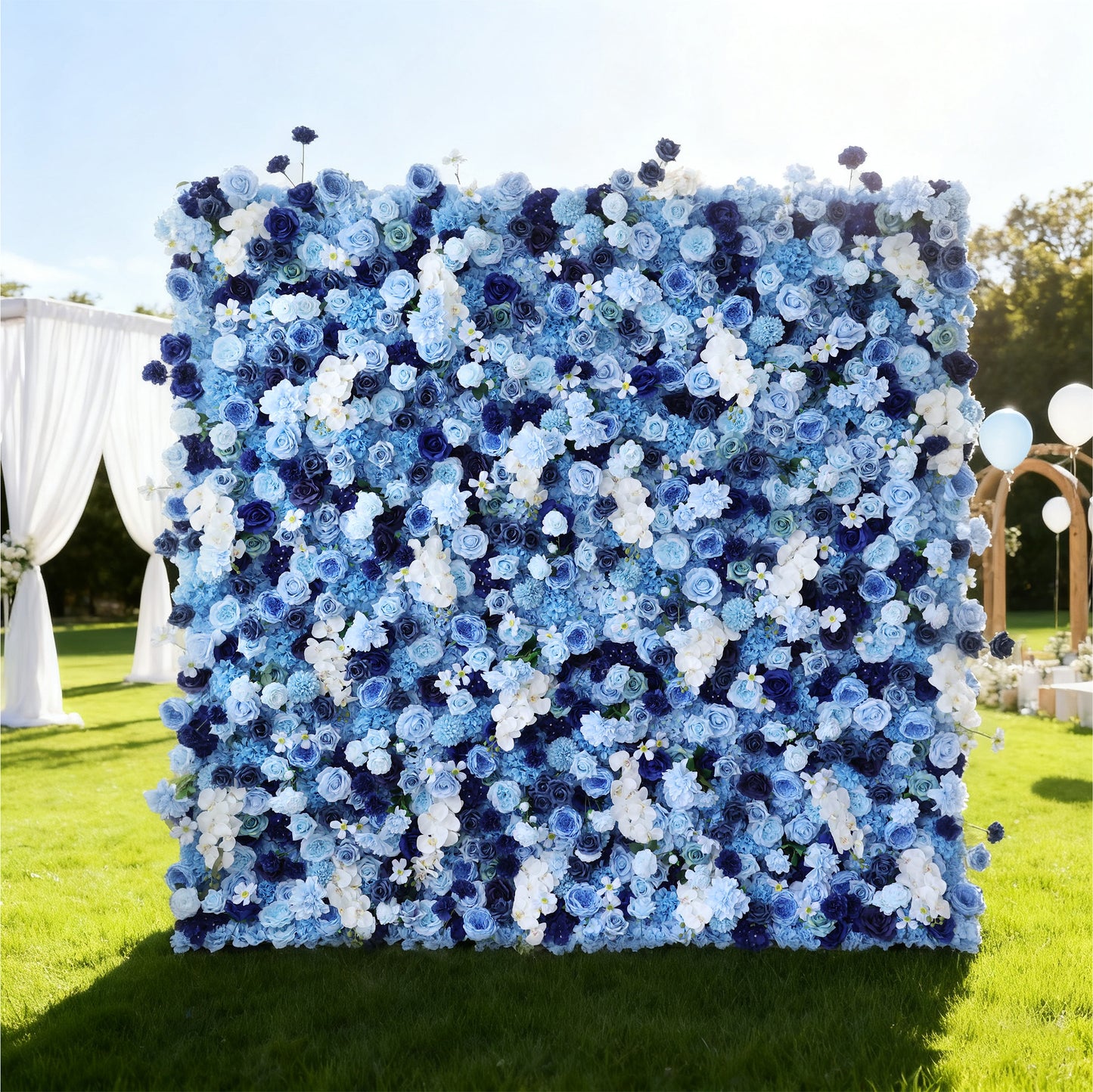 This image features a large, square floral wall outdoors on a lush green lawn. It's densely covered with an array of flowers in various shades of blue and white, creating a serene and elegant display. Bathed in sunlight, with decorative elements like a white curtain and balloons in the background, the scene exudes a sense of natural beauty and refined charm, perfect for events like weddings or celebrations.