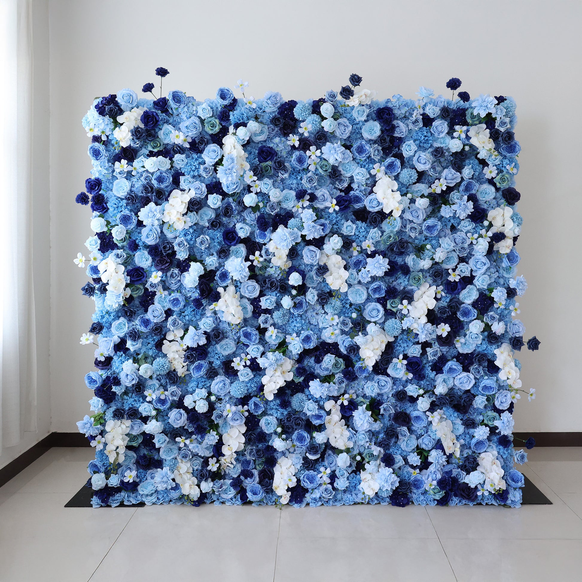 This image showcases a large, dense floral wall indoors. It's covered with an array of artificial flowers in various shades of blue, from deep navy to light sky blue, along with some white blooms. The flowers are densely packed, creating a lush, ocean - like display. Set against a plain white wall and on a light - colored tiled floor, the floral wall exudes a sense of elegance and tranquility, perfect for decorative purposes in events or as a striking interior backdrop.