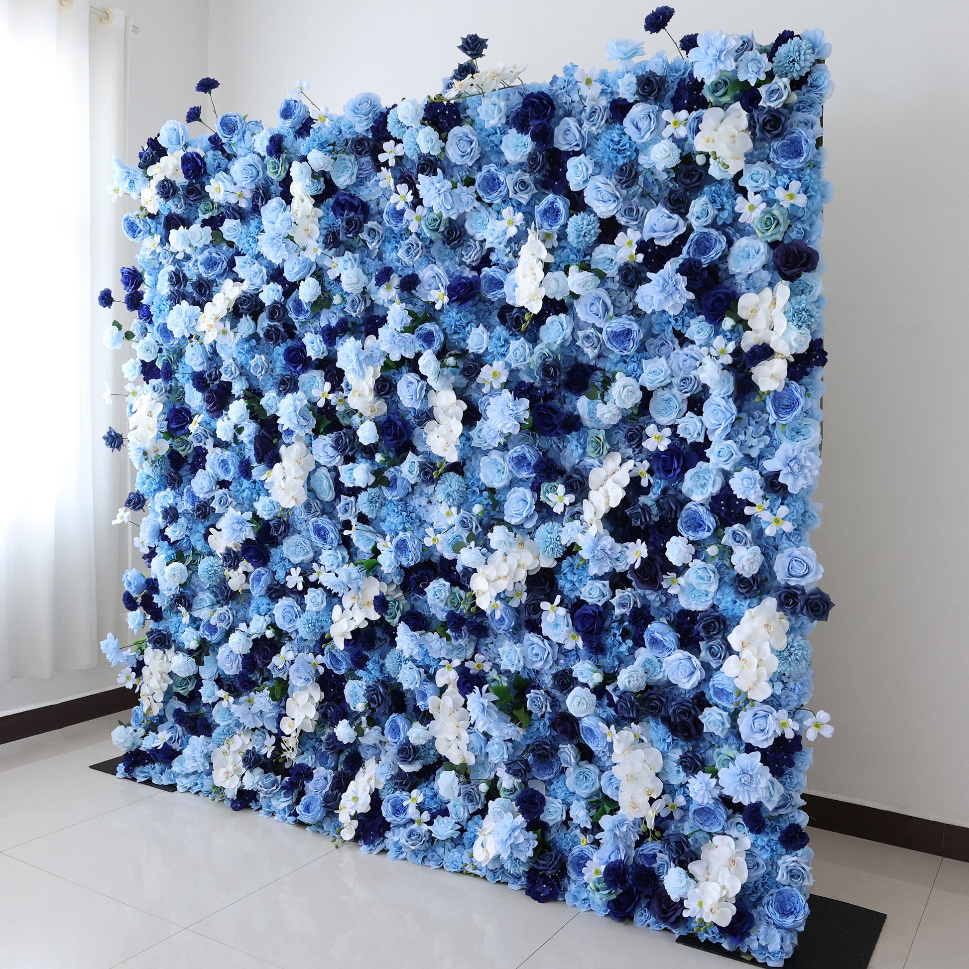 This image showcases a large, dense floral wall indoors. It's covered with an array of flowers in various shades of blue and white, creating a serene and elegant display. Set in a room with white walls and a tiled floor, the harmonious blend of colors and intricate petals exudes a sense of tranquility and refined beauty, perfect for decorative purposes in events or as a striking interior backdrop.