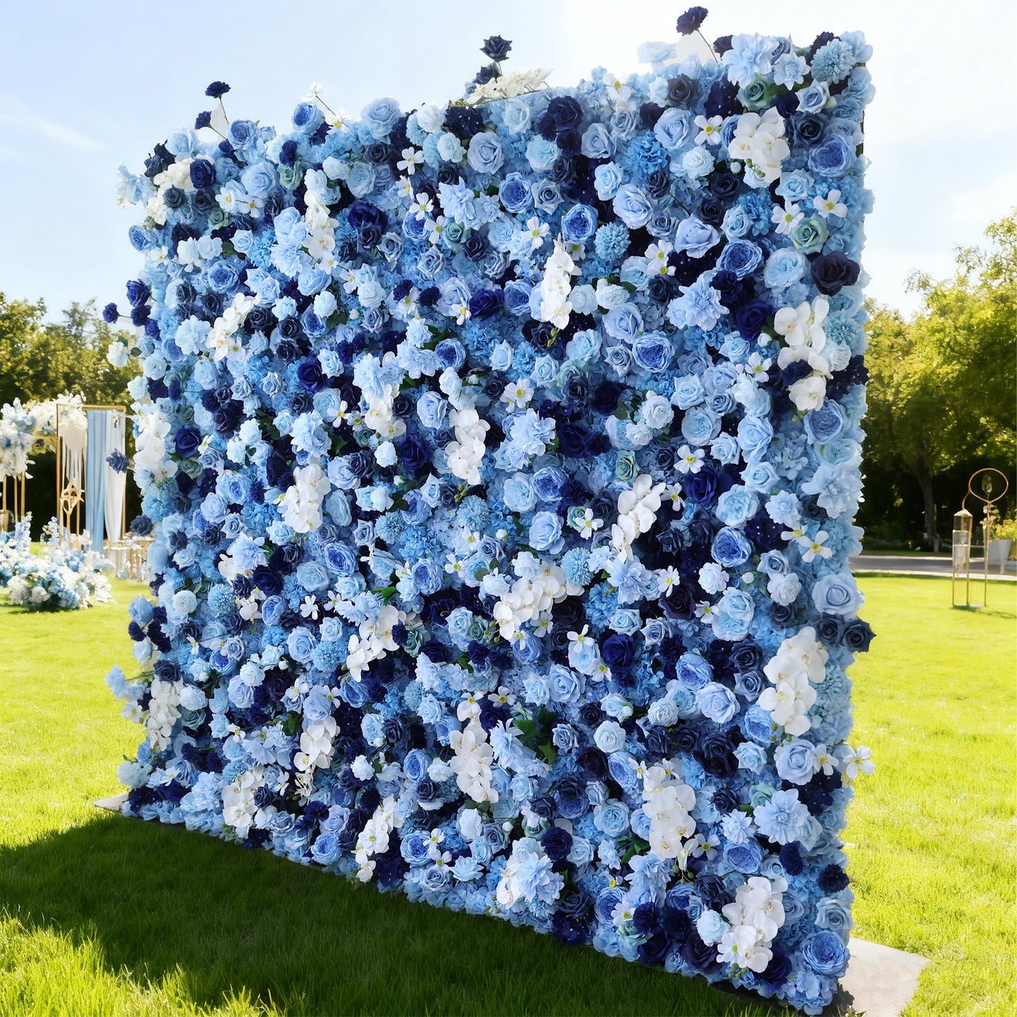 This image features a large, square floral wall outdoors on a lush green lawn. It's densely covered with an array of flowers in various shades of blue and white, creating a serene and elegant display. Bathed in sunlight, with decorative elements and trees in the background, the scene exudes a sense of natural beauty and refined charm, perfect for events like weddings or celebrations.