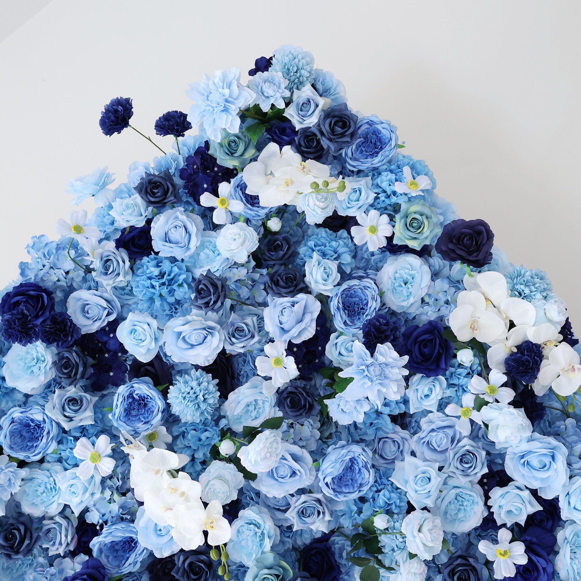 This image showcases a dense, vibrant arrangement of artificial flowers in various shades of blue and white. The flowers, including roses and other blooms, are clustered tightly to form a lush, elegant floral display. The harmonious blend of colors, from light blue to deep navy, along with the intricate petals, creates a serene and refined scene, ideal for decorative purposes in events or as a visually appealing artistic display.