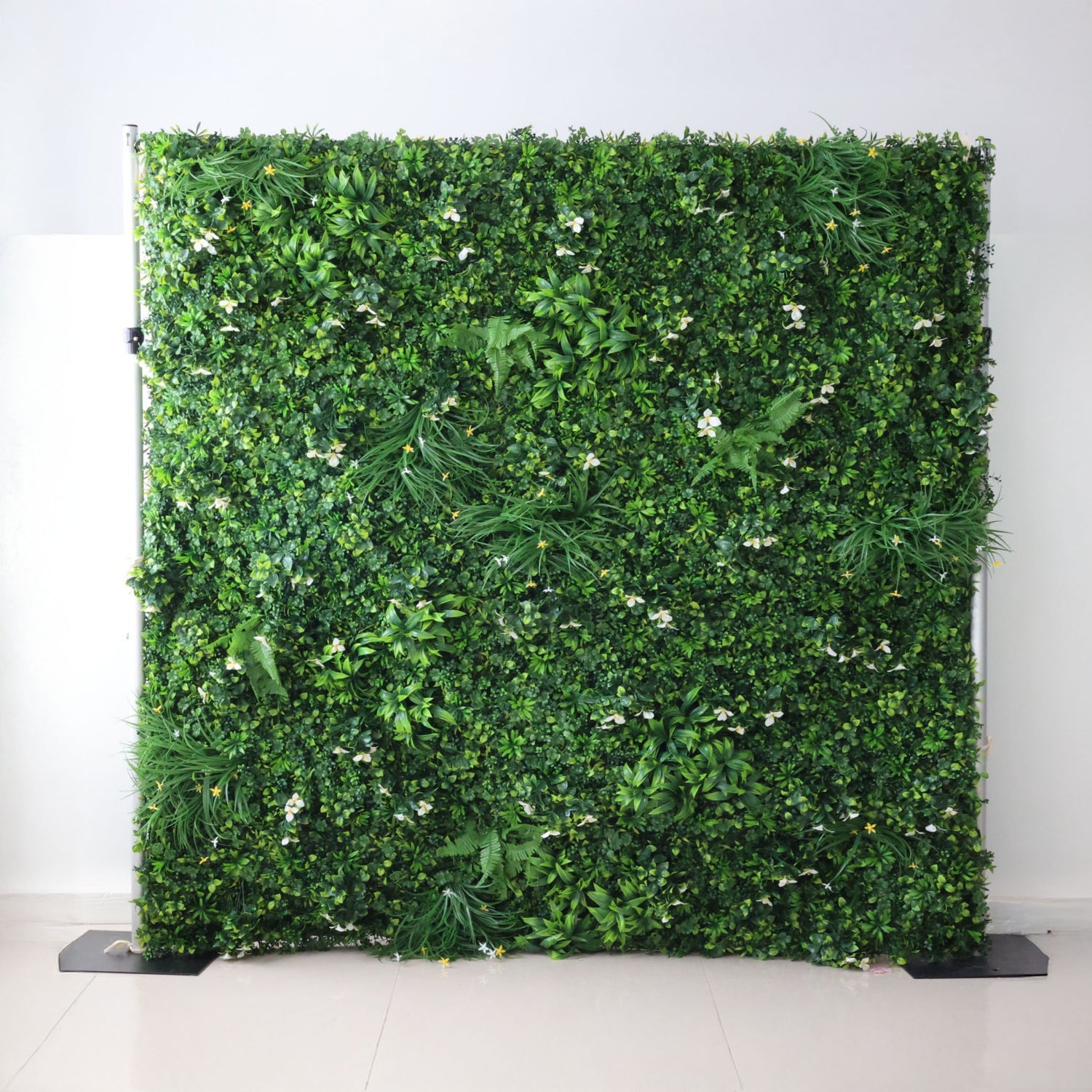 This image showcases a lush artificial greenery wall by ValarFlowers, set indoors against a plain white wall. It features a dense arrangement of varied green foliage, interspersed with delicate white blooms and subtle yellow accents, along with ferns and textured plants. The realistic textures and diverse elements create a rich, natural, and visually captivating display, perfect for adding organic elegance to interiors or events, exuding a serene and sophisticated atmosphere.