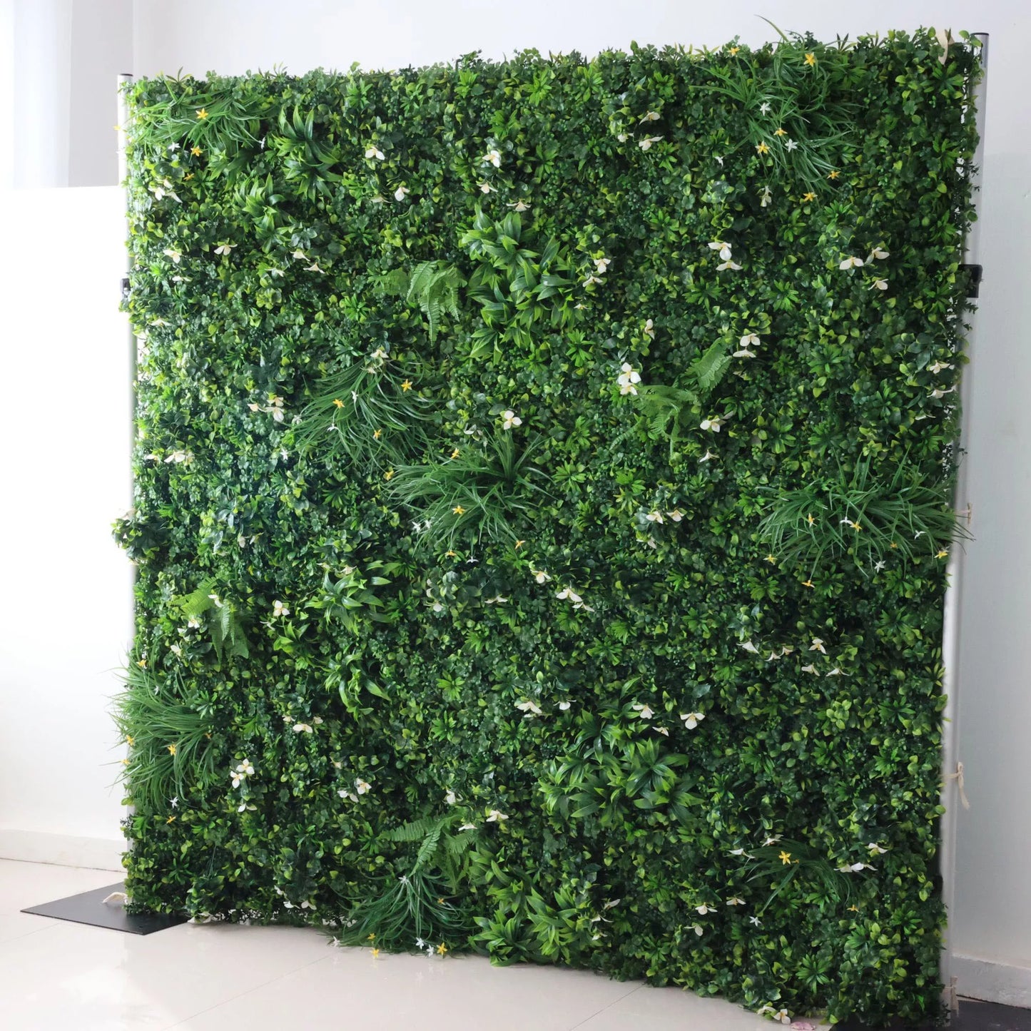 This image showcases a lush artificial greenery wall by ValarFlowers, set indoors. It features a dense arrangement of varied green foliage, interspersed with delicate white blooms and subtle yellow accents, along with ferns and textured plants. The realistic textures and diverse elements create a rich, natural, and visually captivating display, perfect for adding organic elegance to interiors or events, exuding a serene and sophisticated atmosphere.