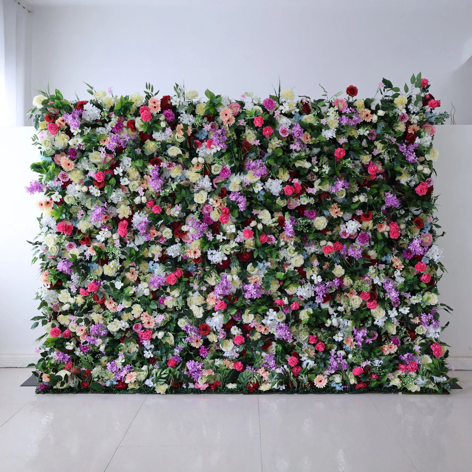 A stunning, floor-to-ceiling floral wall by Valar Flowers against a clean white backdrop bursts with a vibrant rainbow mix: red roses, pink peonies, purple stock, white daisies, yellow blooms, and blue hydrangeas, all lushly packed with greenery. This opulent, full-bloom design is perfect for luxury weddings, photo booths, and upscale event backdrops, creating a breathtaking, festive atmosphere.