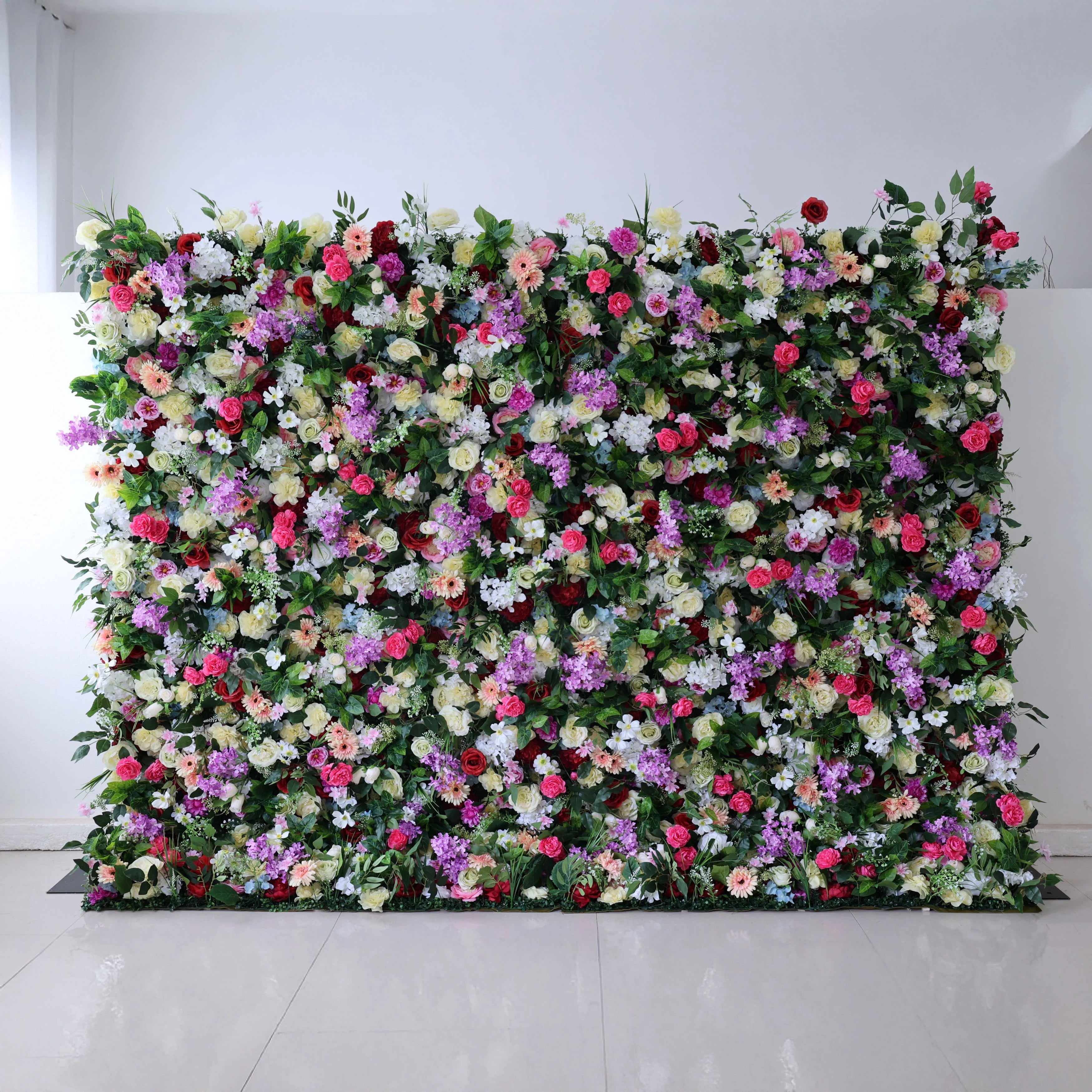A stunning, floor-to-ceiling floral wall by Valar Flowers against a clean white backdrop bursts with a vibrant rainbow mix: red roses, pink peonies, purple stock, white daisies, yellow blooms, and blue hydrangeas, all lushly packed with greenery. This opulent, full-bloom design is perfect for luxury weddings, photo booths, and upscale event backdrops, creating a breathtaking, festive atmosphere.