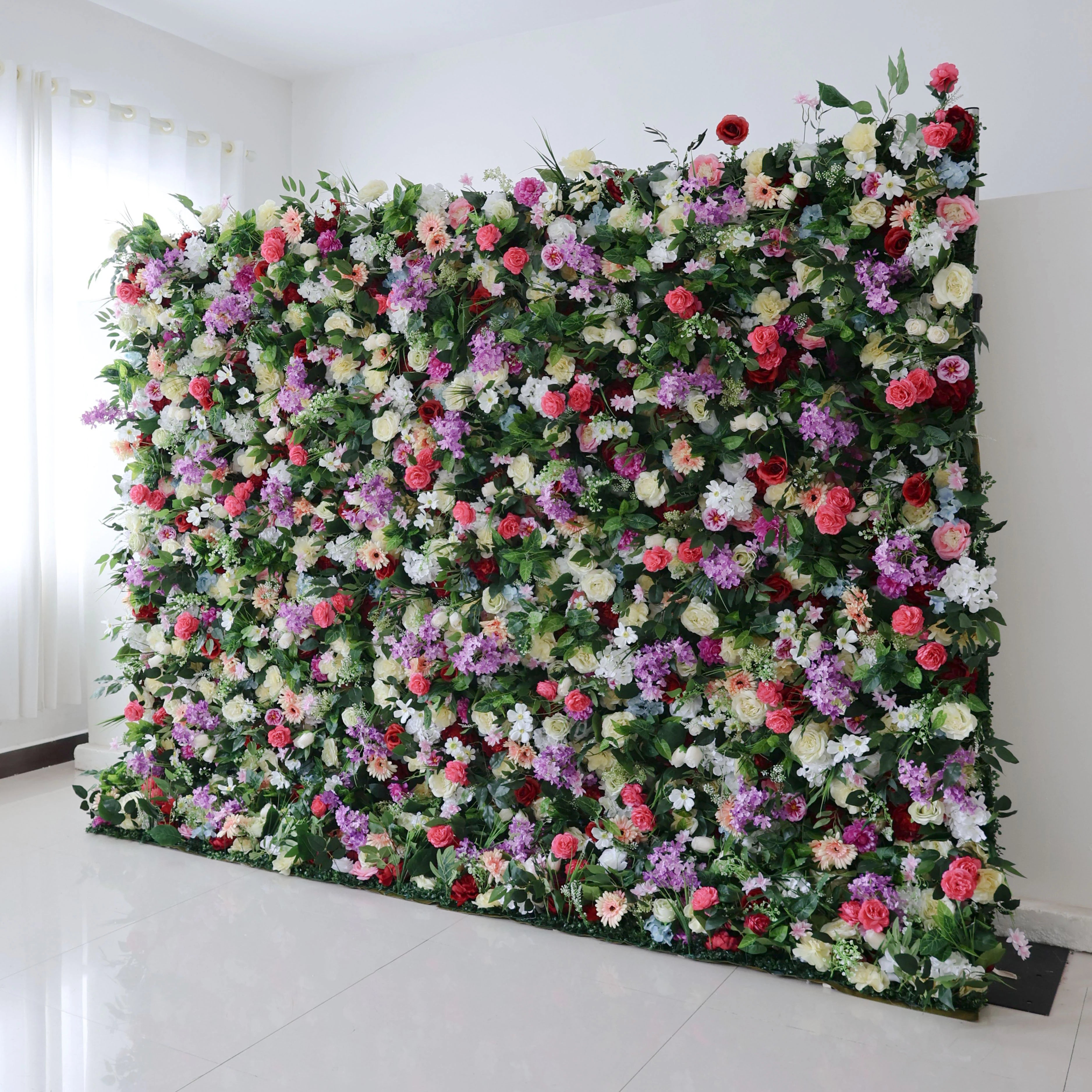 A grand indoor floral wall by Valar Flowers dominates a bright room with white tiled floors and sheer curtains. Densely packed with vivid red roses, soft pink peonies, purple stock, creamy white blooms, and warm peach accents, it’s lushly framed by rich green foliage. This opulent design is ideal for luxury wedding backdrops, bridal shower photo booths, and upscale event decor, exuding elegance and vibrancy.