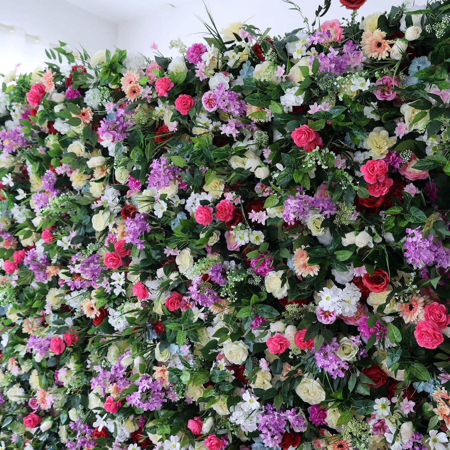 A vibrant, dense floral wall from Valar Flowers bursts with a rich tapestry of blooms: bold red roses, soft pink peonies, deep purple stock, creamy white carnations, sunny yellow gerberas, and delicate blue hydrangeas, all nestled in lush green foliage. This eye-catching, full-bloom design delivers a cheerful, festive aesthetic, perfect for wedding backdrops, photo booths, birthday parties, or vibrant event decor.