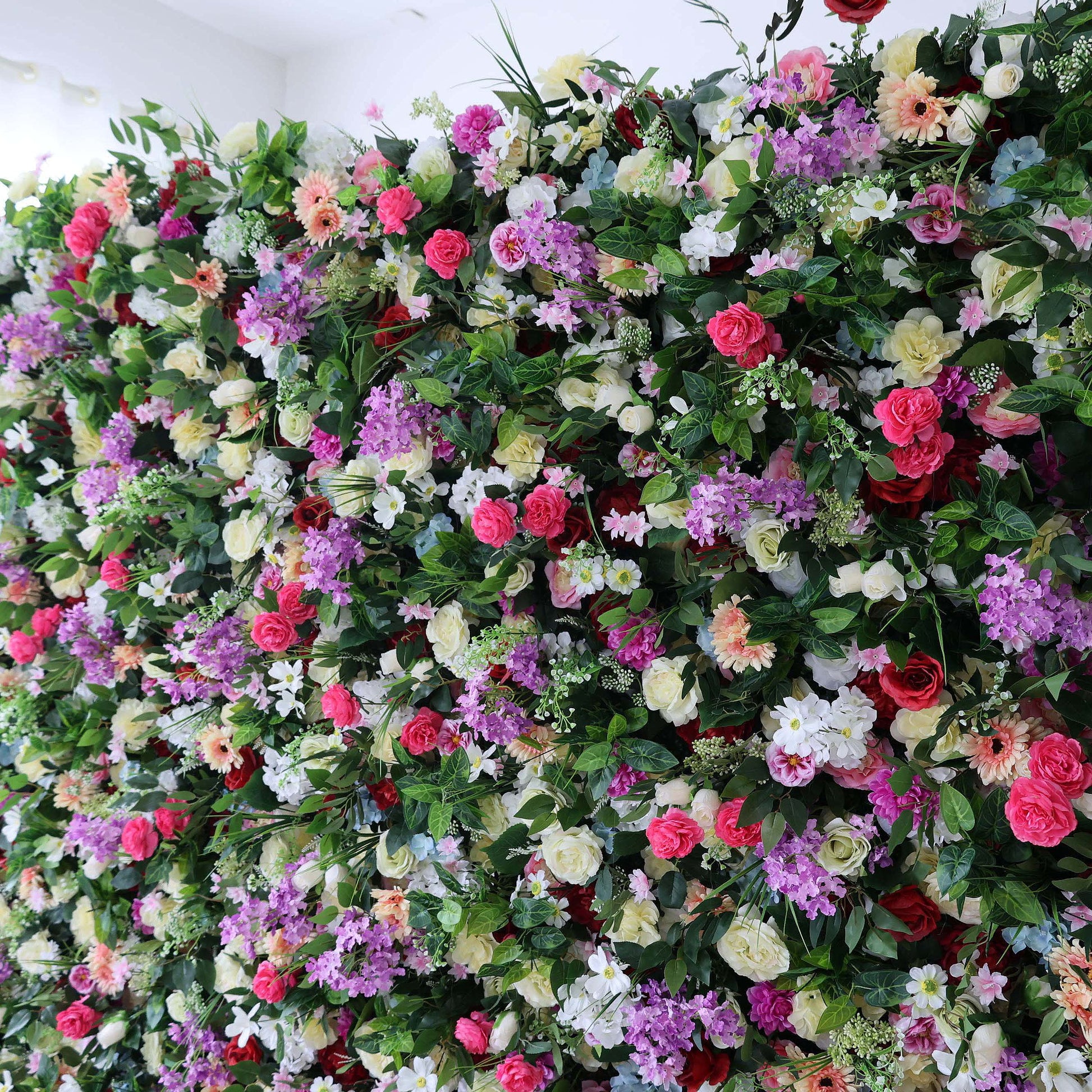 A vibrant, dense floral wall from Valar Flowers bursts with a rich tapestry of blooms: bold red roses, soft pink peonies, deep purple stock, creamy white carnations, sunny yellow gerberas, and delicate blue hydrangeas, all nestled in lush green foliage. This eye-catching, full-bloom design delivers a cheerful, festive aesthetic, perfect for wedding backdrops, photo booths, birthday parties, or vibrant event decor.
