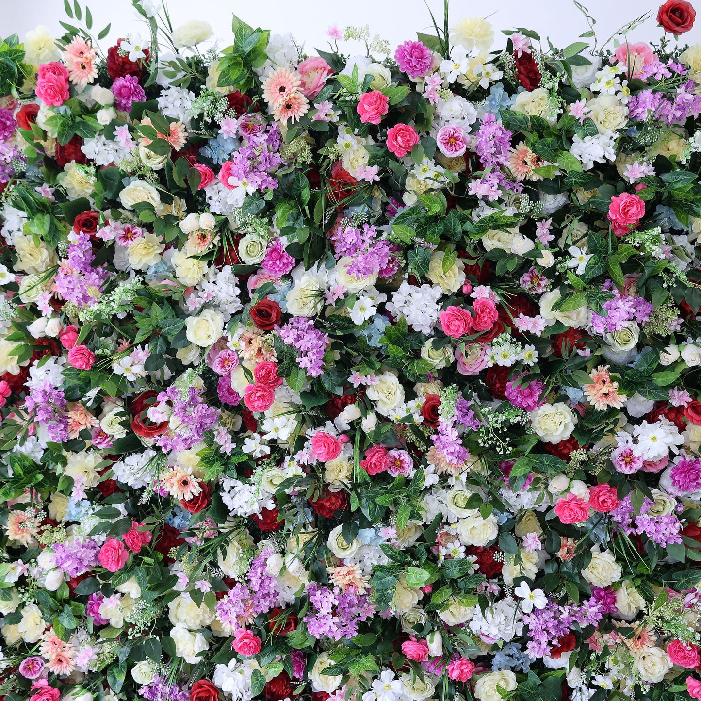 A vibrant, dense floral wall from Valar Flowers bursts with a rainbow of blooms—bold red roses, soft pink peonies, purple stock, creamy white carnations, and sunny yellow accents—nestled in lush green foliage. This eye-catching, full-bloom design delivers a cheerful, festive aesthetic, perfect for party backdrops, photo booths, or vibrant event decor.