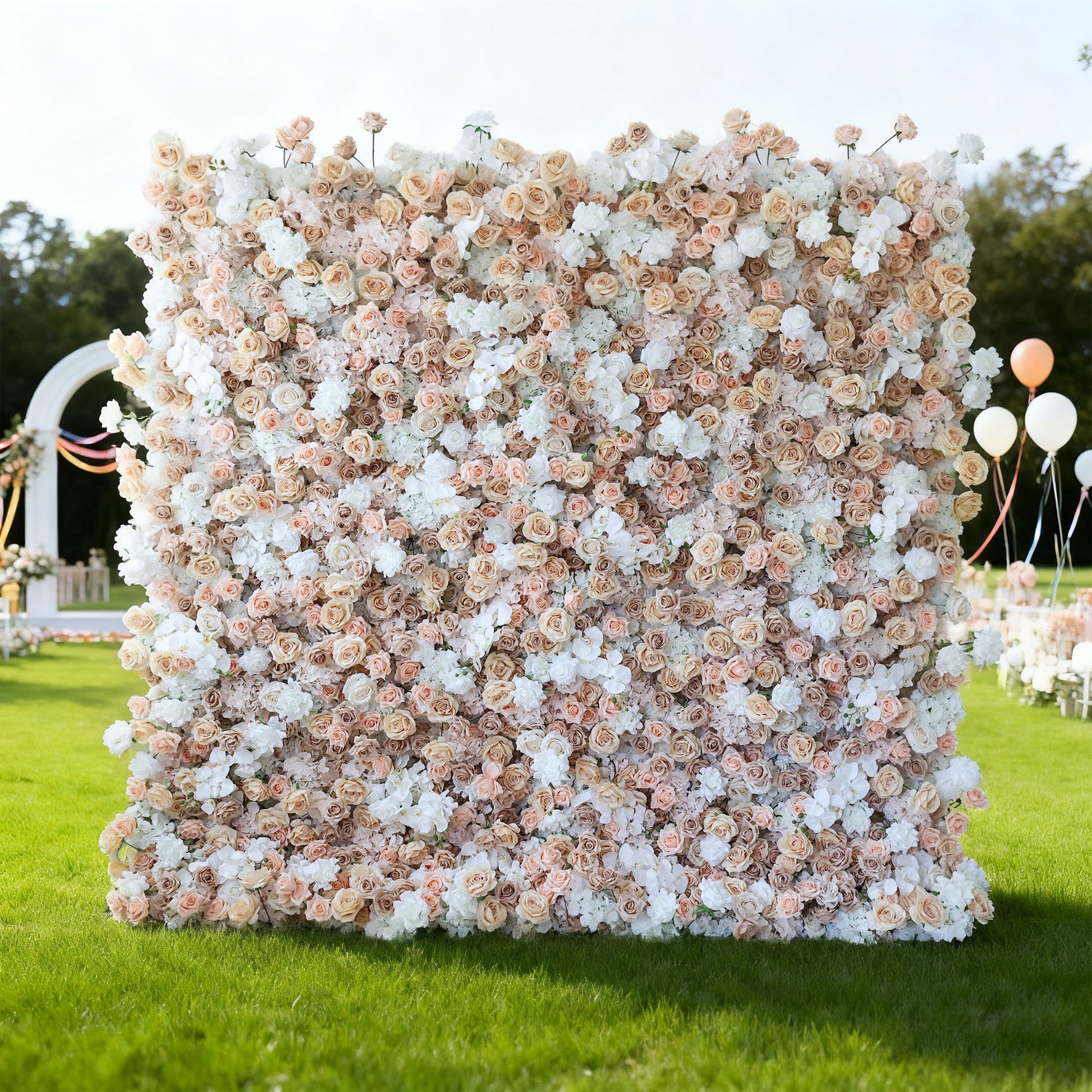 This image features a large, square floral wall outdoors on a lush green lawn. It's densely covered with an array of flowers in soft hues of beige, white, and light pink, creating a romantic and elegant display. Bathed in sunlight, with trees and a clear sky in the background, and accompanied by decorative elements like balloons and an arch, the scene exudes a sense of natural beauty and charm, perfect for events like weddings or celebrations.