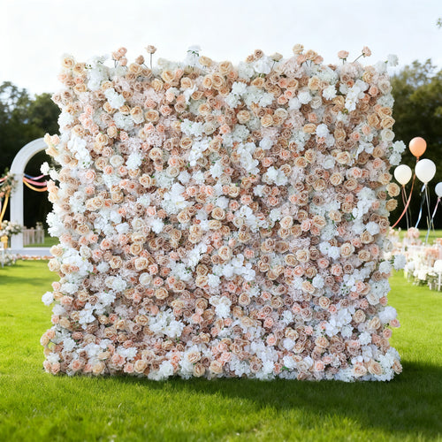This image features a large, square floral wall outdoors on a lush green lawn. It's densely covered with an array of flowers in soft hues of beige, white, and light pink, creating a romantic and elegant display. Bathed in sunlight, with trees and a clear sky in the background, and accompanied by decorative elements like balloons and an arch, the scene exudes a sense of natural beauty and charm, perfect for events like weddings or celebrations.
