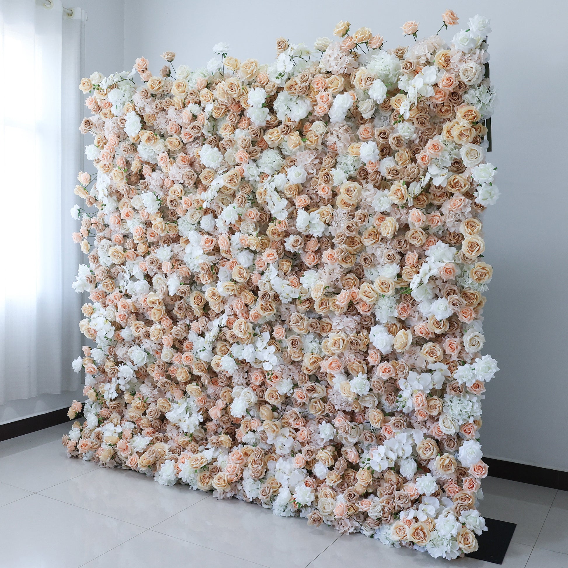 This image showcases a large, dense floral wall indoors. It's covered with an array of flowers in soft hues of beige, white, and light pink, creating a romantic and elegant display. Set against a plain wall with a tiled floor, the scene exudes a sense of sophistication and charm, perfect for decorative purposes in events or as a striking interior backdrop.