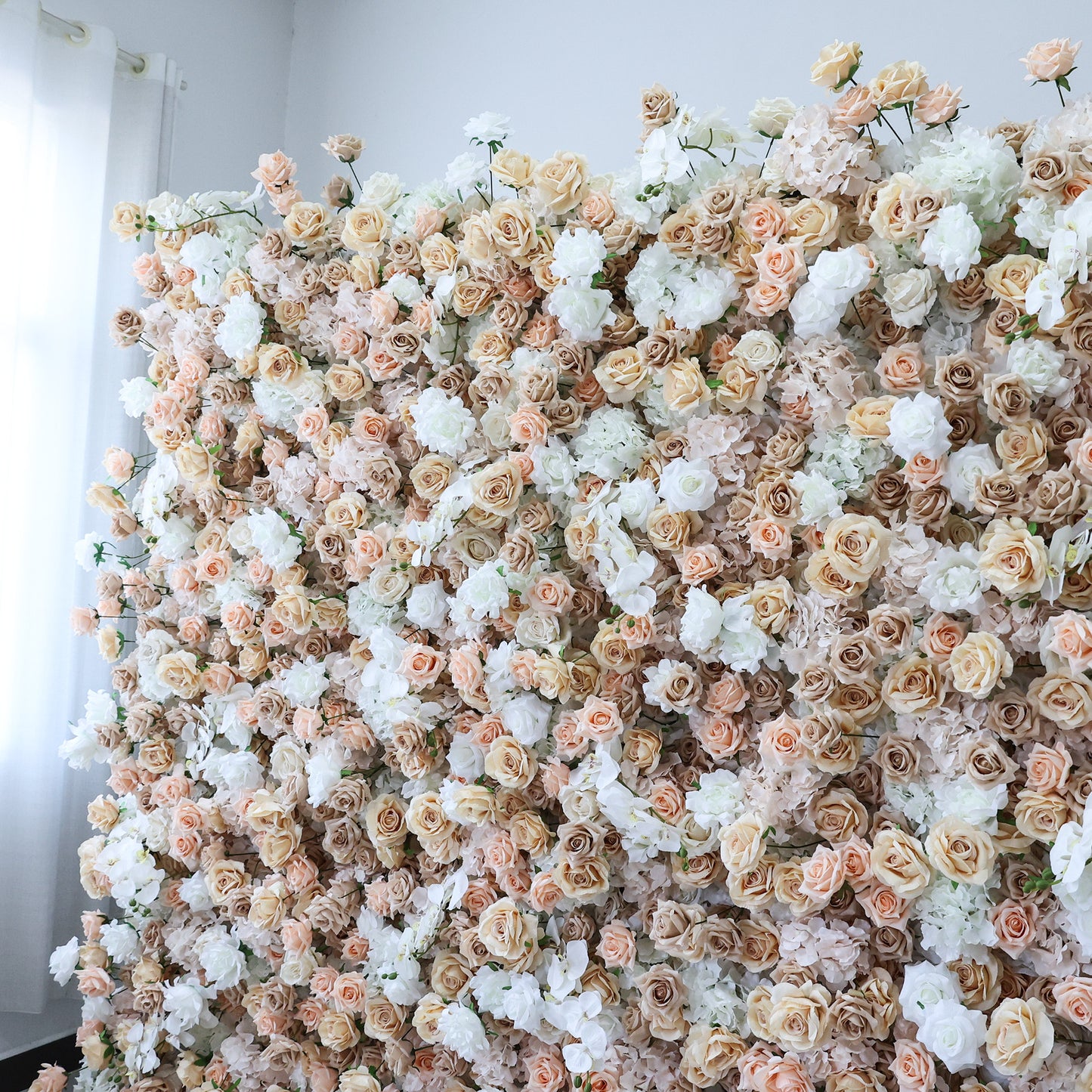 This image displays a large, dense floral wall indoors. It's covered with an array of flowers in soft hues of beige, white, and light pink, creating a romantic and elegant display. Set in a room with white walls and a curtain, the scene exudes a sense of sophistication and charm, perfect for decorative purposes in events or as a striking interior backdrop.