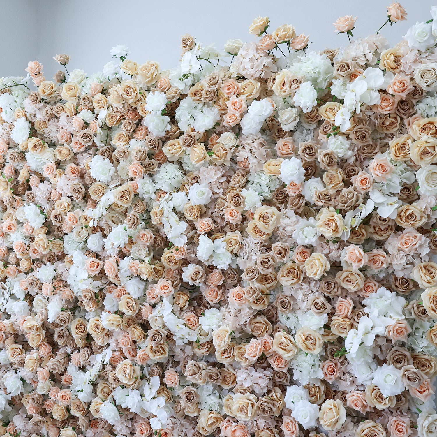 This image presents a dense, vibrant arrangement of artificial flowers in soft hues of beige, white, and light pink. The flowers, including roses and other blooms, are clustered tightly to form a lush, romantic floral tapestry. Set against a plain backdrop, the intricate petals and harmonious color palette create an elegant, charming scene, ideal for event decor or as a visually appealing artistic display.