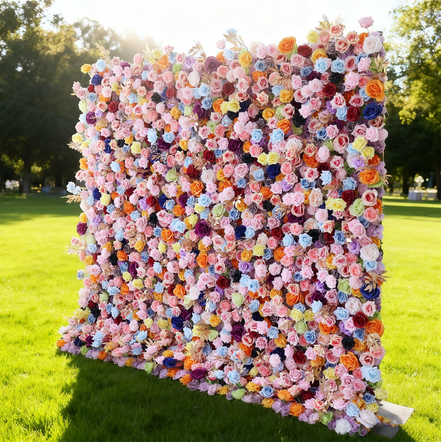 This image features a large, square floral wall outdoors on a lush green lawn. It's densely covered with an array of vibrant flowers in a wide spectrum of colors, including pink, purple, blue, orange, and yellow, creating a lively and eye-catching display. Bathed in sunlight, with trees and a clear sky in the background, the scene exudes a sense of natural beauty and festive charm, perfect for events or as a stunning decorative backdrop.