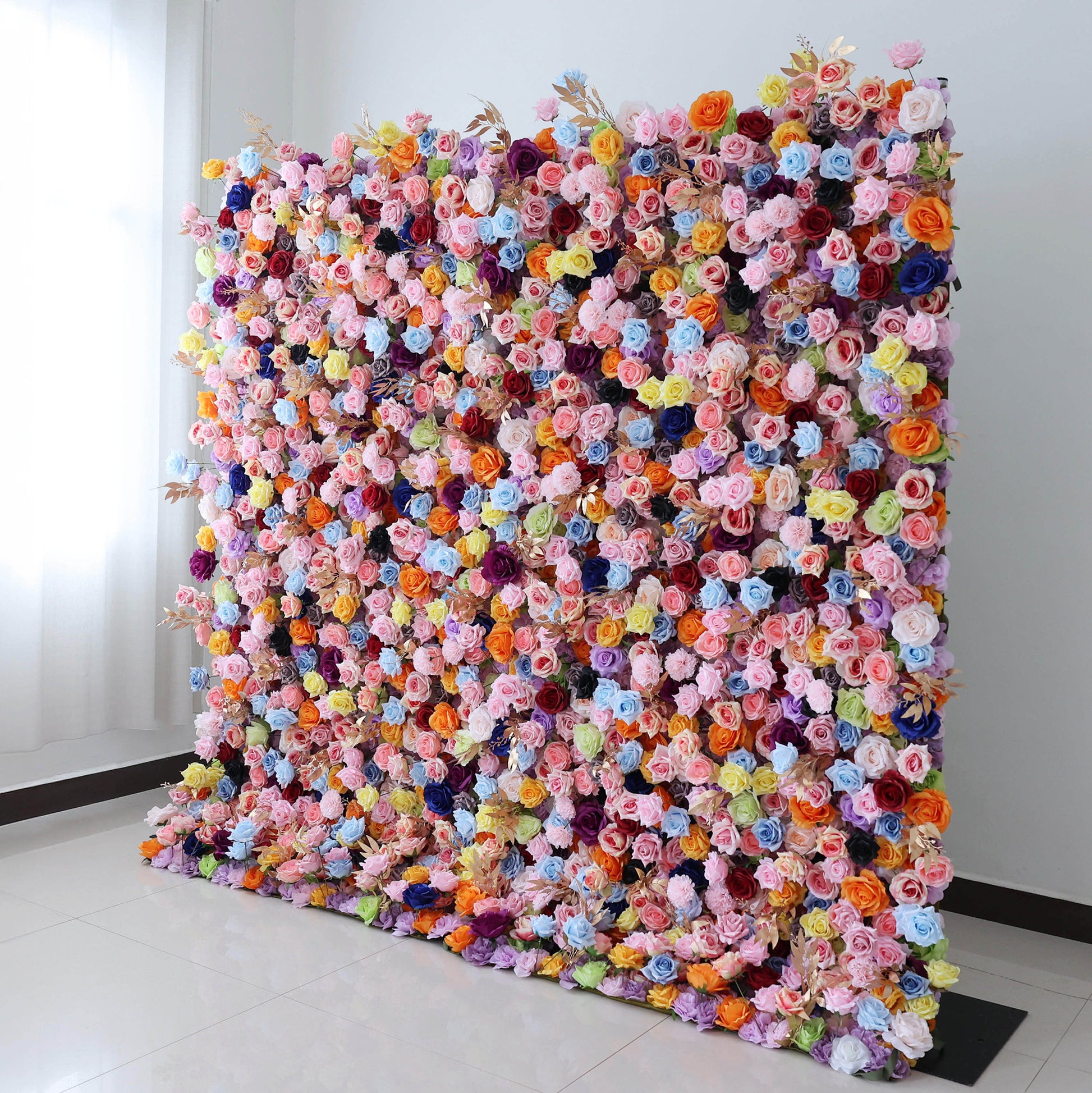 This image showcases a large, dense floral wall indoors. It's covered with an array of artificial flowers in vibrant colors like pink, blue, orange, yellow, and purple. The flowers, including roses and other blooms, are densely packed, creating a lush, colorful display. Set against a plain white wall and on a light - colored tiled floor, the floral wall exudes a sense of natural beauty and charm, perfect for decorative purposes in events or as a striking interior backdrop.