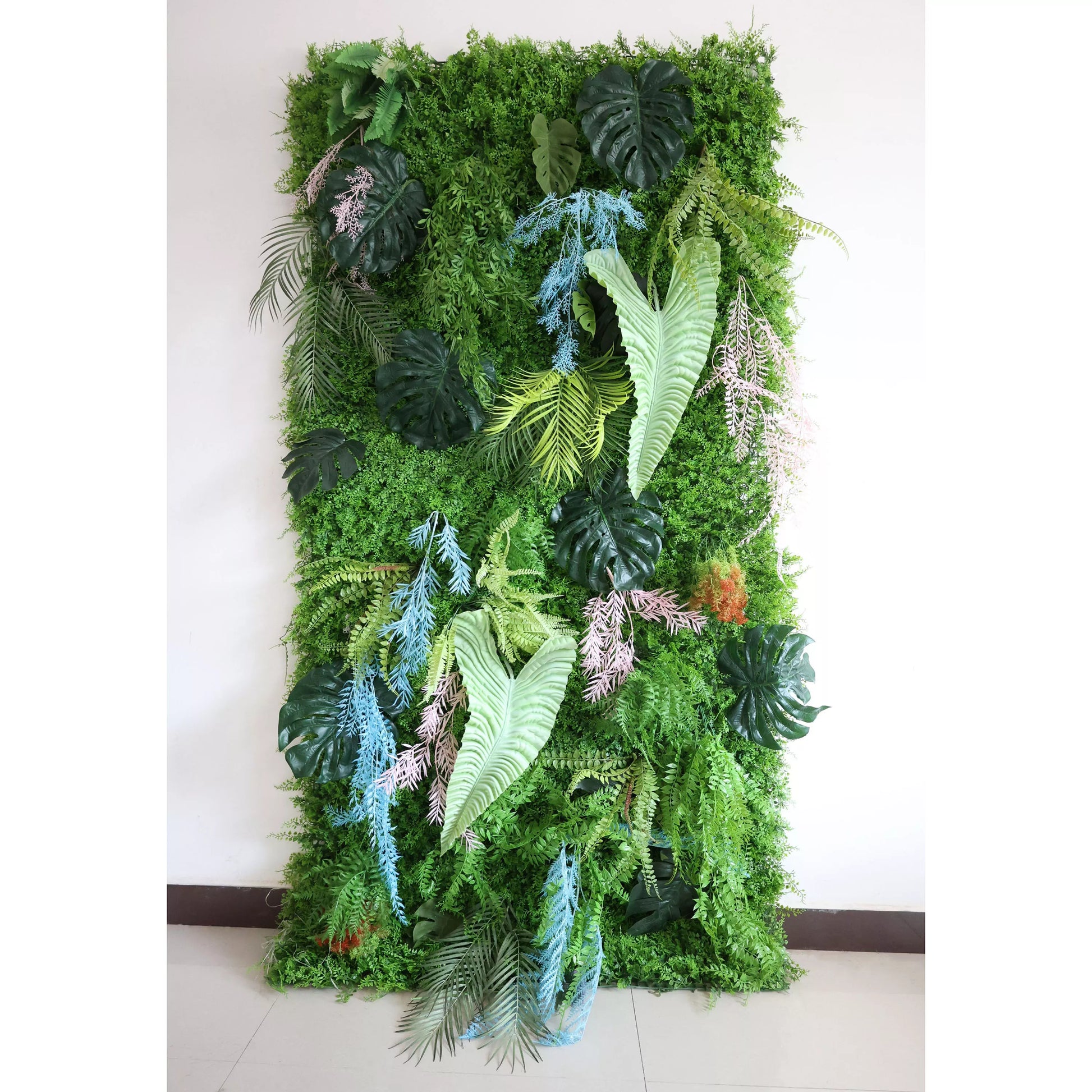 Palm-like leaves add a tropical touch. Among the greenery, non-green foliage offers unique color pops: light blue, pinkish-lavender artificial plants (likely ornamental grasses) weave through, contrasting the dominant green. Orange-tinged elements, mimicking flowering or autumnal plants, are also present. This vibrant, low-maintenance green wall serves as a natural backdrop, ideal for interiors or events.