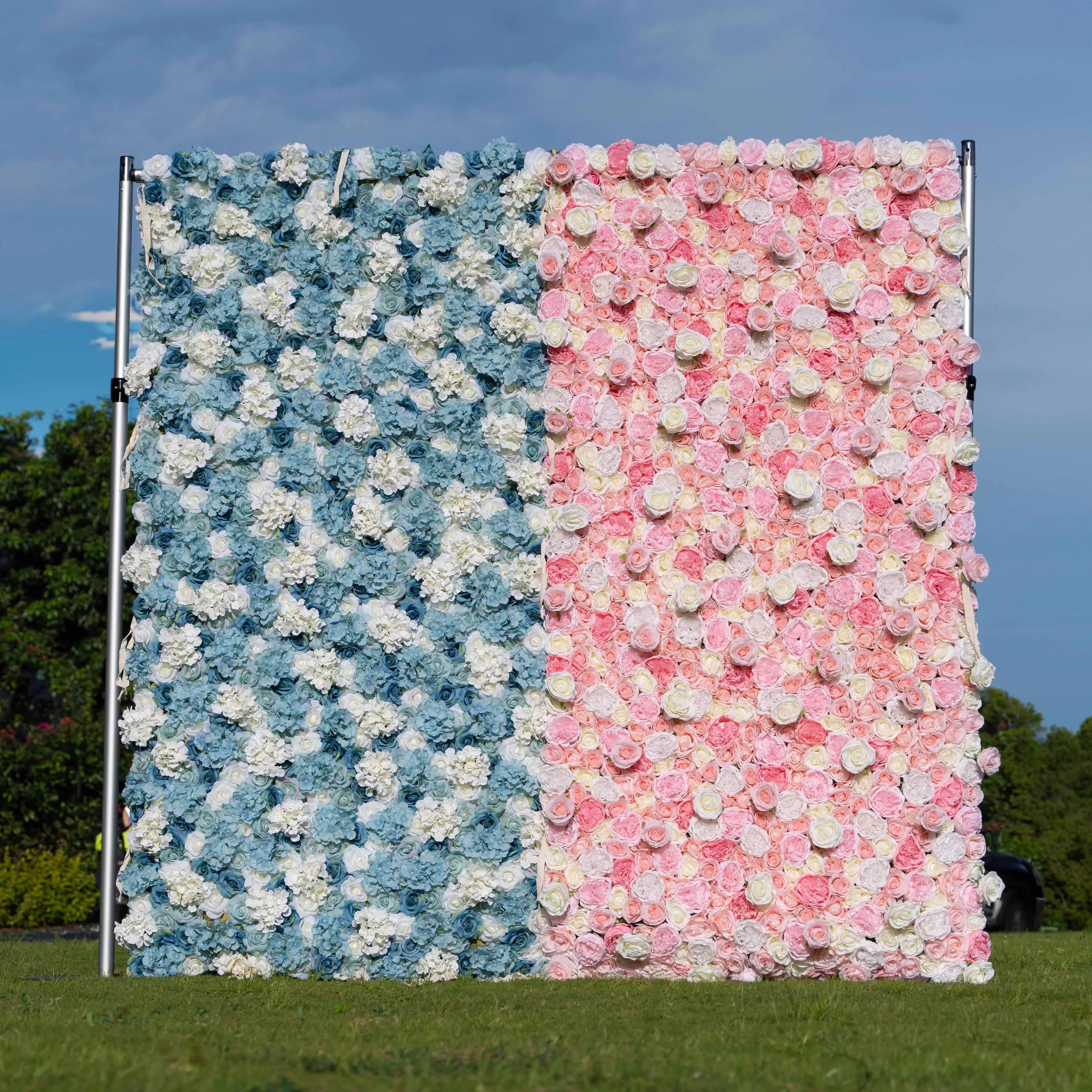 An outdoor floral wall on a metal frame divides into two sections. One side features blue and white artificial flowers, while the other has pink and white ones, set against a grassy area with a sky backdrop. 