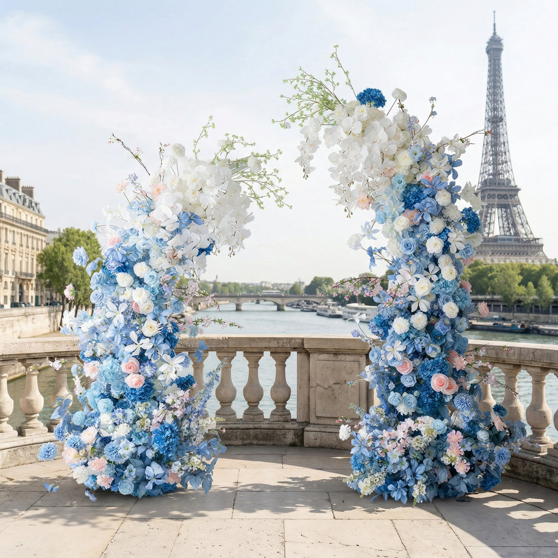 A stunning, asymmetrical floral arch from Valar Flowers, featuring a serene palette of sky blue, navy, and creamy white blooms—including hydrangeas, roses, and delicate orchids—with subtle blush pink accents. Set against the iconic Eiffel Tower and Seine River in Paris, it creates a sophisticated, romantic atmosphere perfect for a luxury wedding or special celebration.