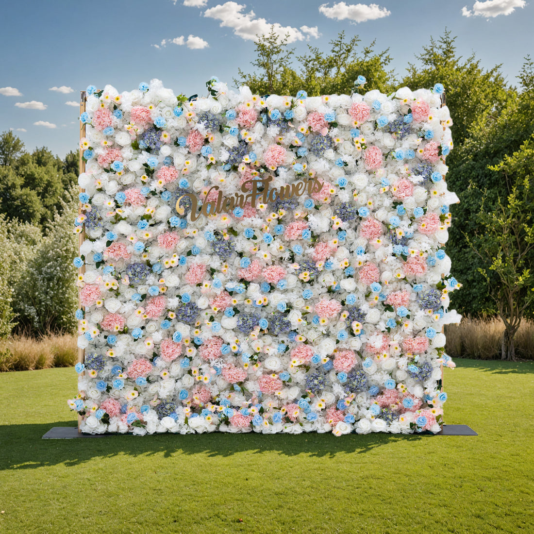 This photo showcases a magnificent floral backdrop from Valar Flowers, set against a backdrop of lush greenery and a bright blue sky with scattered clouds. The backdrop is a dense arrangement of artificial flowers in various shades of pink, blue, and white, creating a visually stunning and vibrant display. The gold lettering of &quot;ValarFlowers&quot; adds a touch of elegance and sophistication, making it an ideal decorative piece for weddings, photo shoots, or other special events.