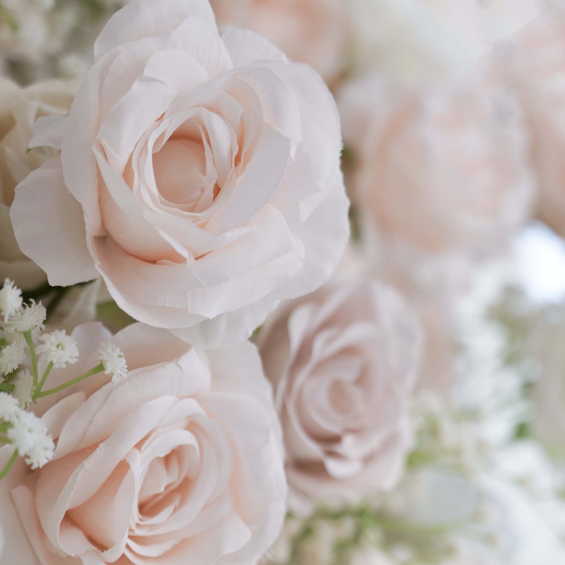 This close - up image highlights a section of a floral backdrop. The flowers predominantly include artificial roses in soft, pastel shades such as light pink and off - white. Delicate sprigs of baby's - breath - like greenery are interspersed among the roses. The overall color palette is gentle and soothing, creating a romantic and elegant style, perfect for wedding or event decorations. 
