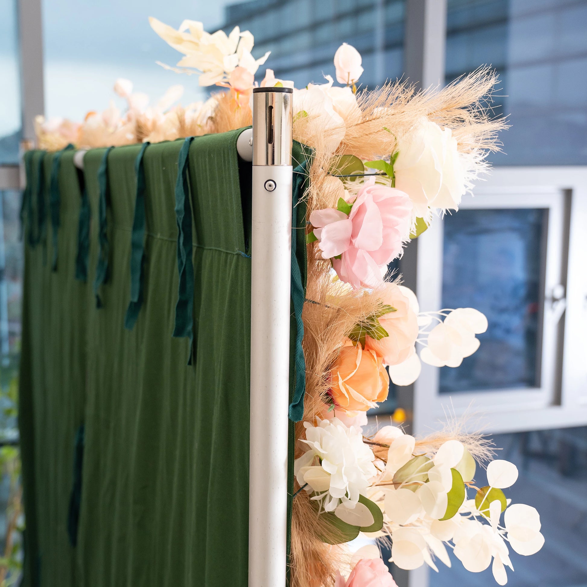 This photo captures a section of a decorative setup, featuring a metallic pole adorned with an arrangement of artificial flowers in soft pink, peach, and white hues, along with fluffy pampas grass. The pole is part of a structure draped with rich green fabric that has hanging green ribbons. Ideal for events like weddings or upscale parties, it adds an elegant, romantic touch to the decor. 
