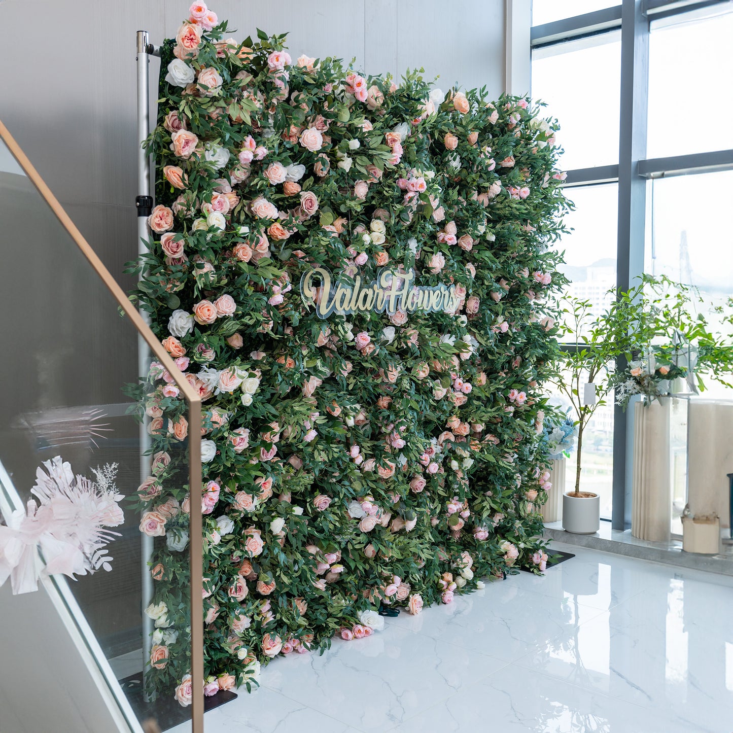 This photo showcases a large, lush floral backdrop indoors, densely adorned with pink and white roses interspersed among green foliage. The center features the text "ValarFlowers" in elegant golden lettering. Ideal for events like corporate galas or a "Floral Elegance Affair", it creates a sophisticated and inviting atmosphere, perfect for upscale indoor celebrations. 