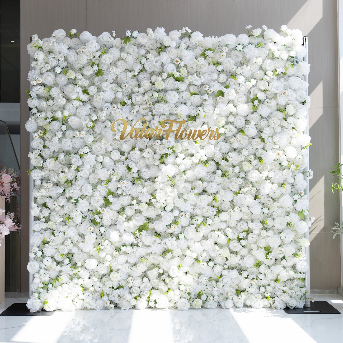 This floral backdrop is densely packed with artificial white roses and small - bud flowers, interspersed with green leaves. The all - white color scheme, accented by the greenery, creates a clean, elegant, and pure aesthetic. The golden “ValarFlowers” signage adds a touch of sophistication. The overall style is classic and refined, suitable for formal events. 
