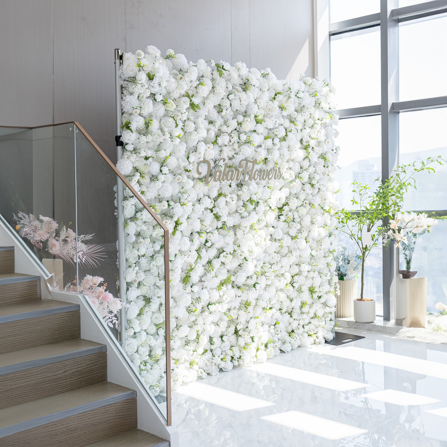 This floral backdrop is densely covered with artificial white flowers, mainly roses and some other assorted blooms, interspersed with green leaves. The all - white color palette, complemented by the greenery, creates a clean, elegant, and sophisticated style. The golden “ValarFlowers” sign adds a touch of luxury, making it perfect for formal events. 