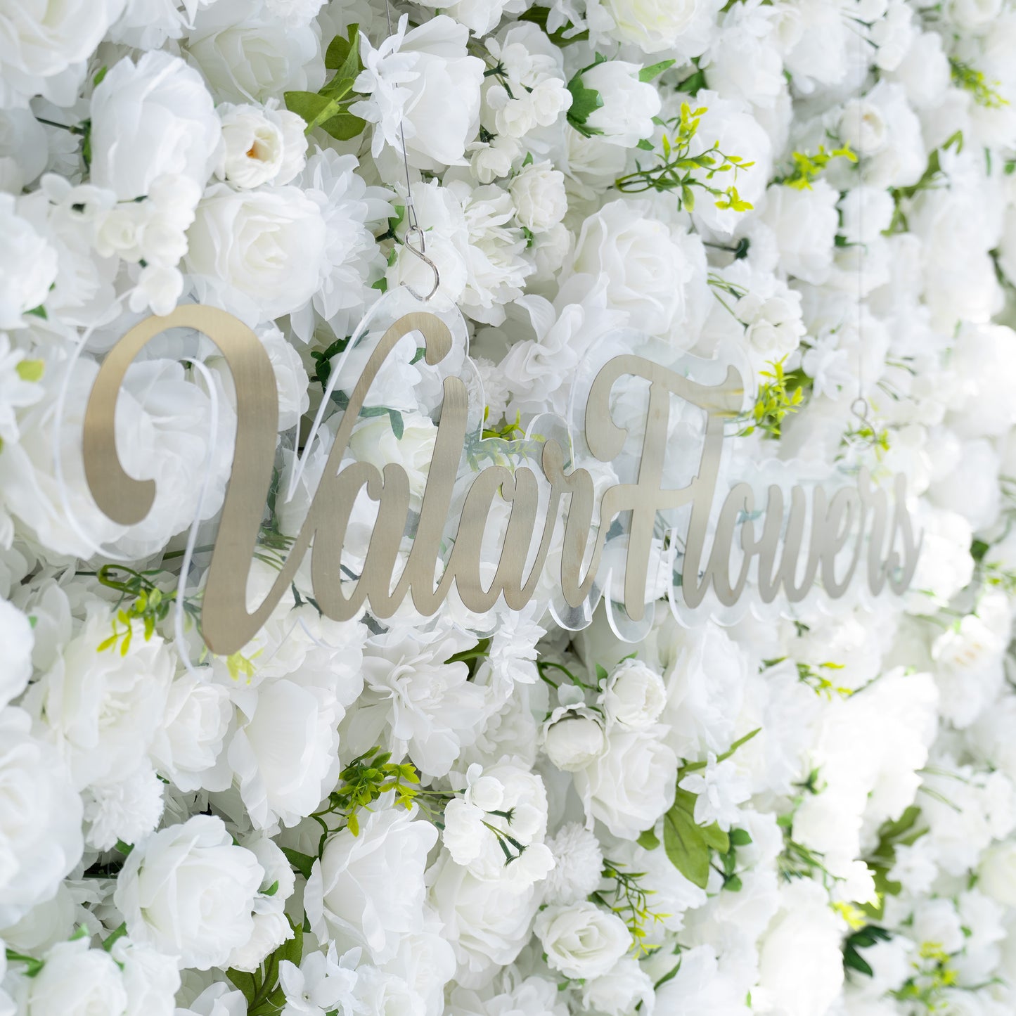 This backdrop features a dense arrangement of artificial white flowers, including roses and various other blooms, interspersed with green leaves. The all - white color scheme, accented by the greenery, exudes a clean and elegant style. The addition of the golden “ValarFlowers” signage enhances the sophisticated and refined aesthetic, making it suitable for formal events. 