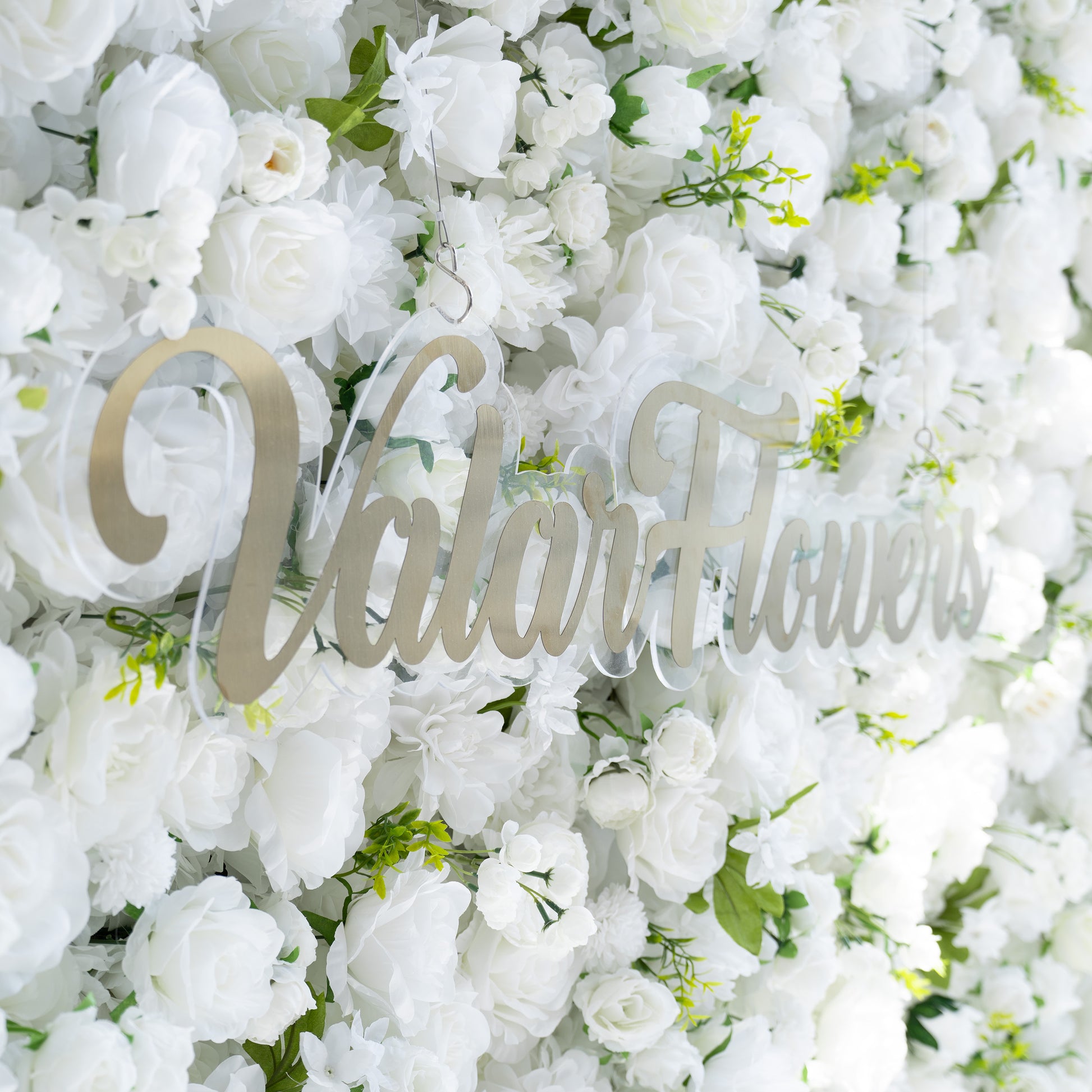 This backdrop features a dense arrangement of artificial white flowers, including roses and various other blooms, interspersed with green leaves. The all - white color scheme, accented by the greenery, exudes a clean and elegant style. The addition of the golden “ValarFlowers” signage enhances the sophisticated and refined aesthetic, making it suitable for formal events. 