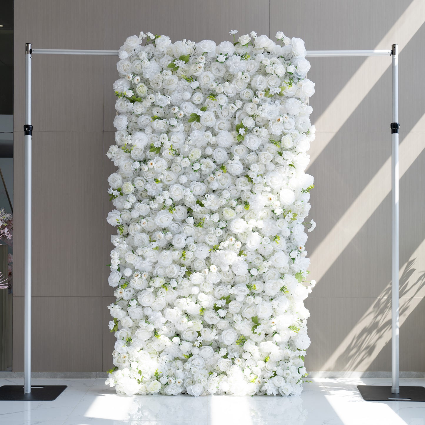 This floral backdrop is supported by a metal frame and densely adorned with artificial white flowers, mainly roses and some smaller - bud blooms, interspersed with green leaves. The all - white color scheme, accented by greenery, creates a clean, elegant, and sophisticated style, perfect for formal events like weddings or corporate gatherings. 