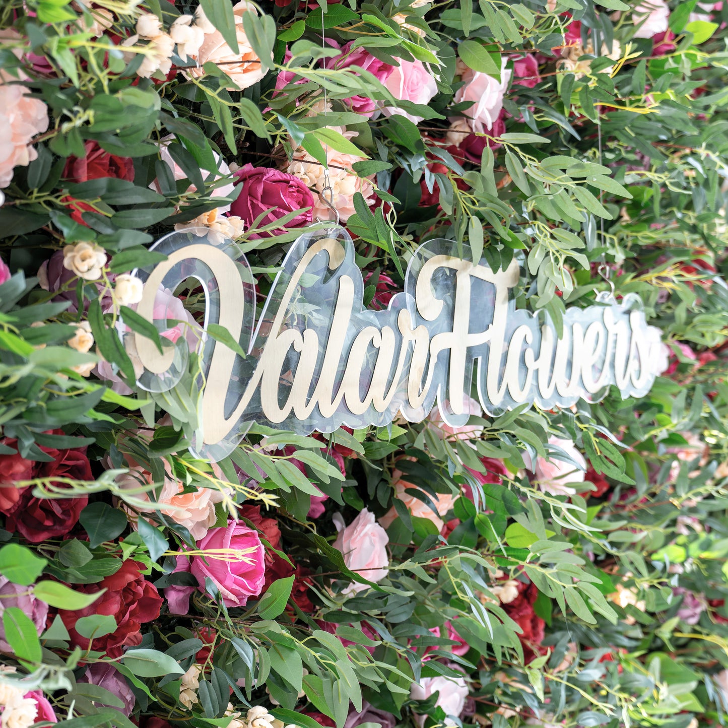 A close - up of a clear acrylic sign with gold “ValarFlowers” script, nestled in a lush artificial flower wall. The wall features pink, red, and peach roses, plus green foliage, creating a vibrant, decorative backdrop that highlights the branding element. 